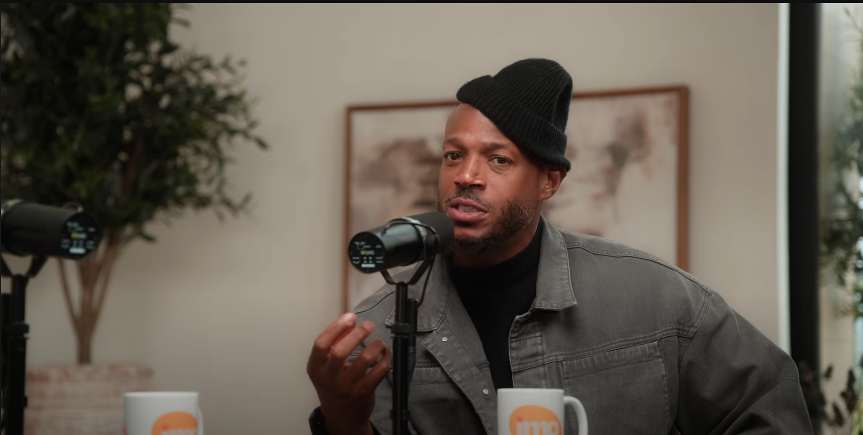 Person speaking into a microphone during a podcast interview, wearing a beanie and casual jacket, with two branded mugs on the table