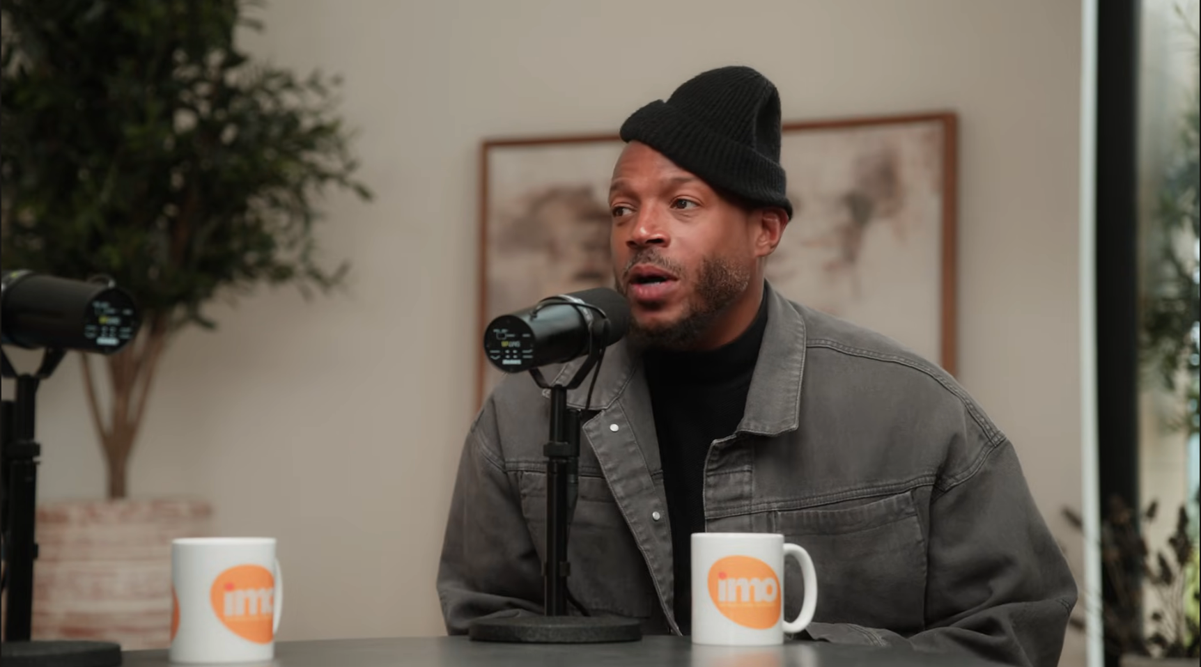 Person wearing a black beanie and denim jacket speaking into a microphone in a casual setting, with two coffee mugs on the table