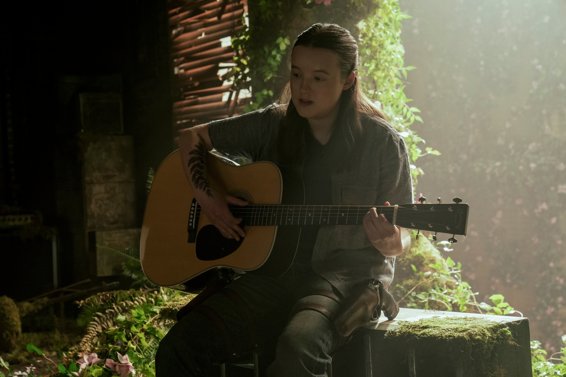 Persona tocando guitarra acústica en un escenario natural con vegetación de fondo. Lleva ropa casual y tiene el cabello suelto