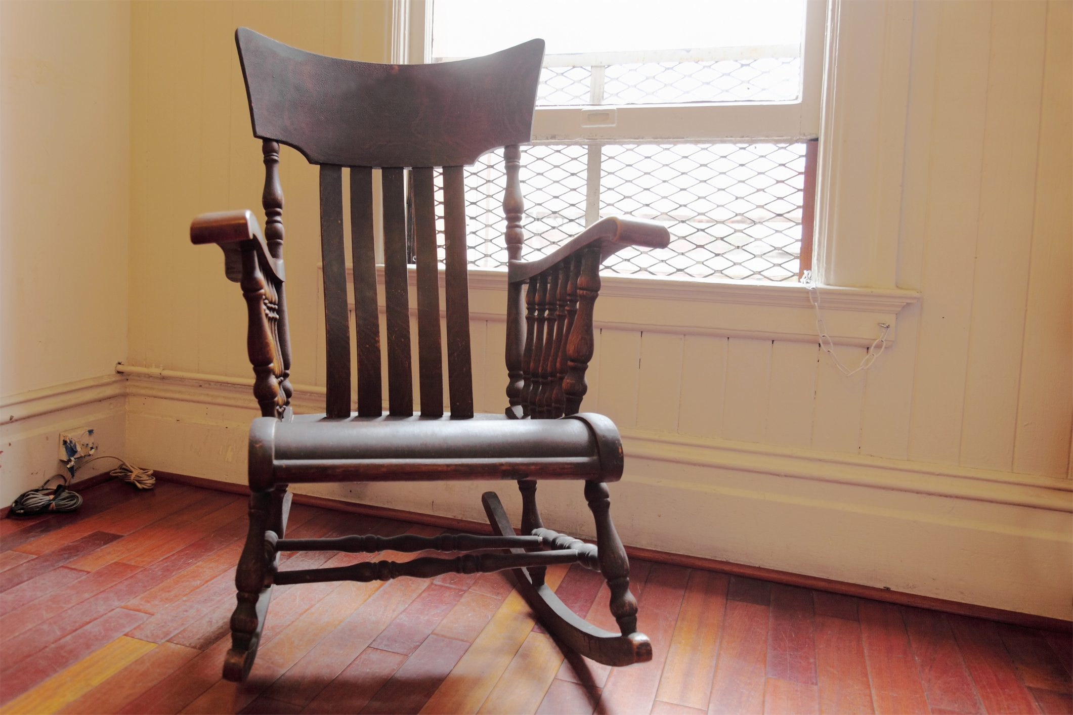 A vintage wooden rocking chair sits in a room with hardwood floors, next to a large window with a mesh screen