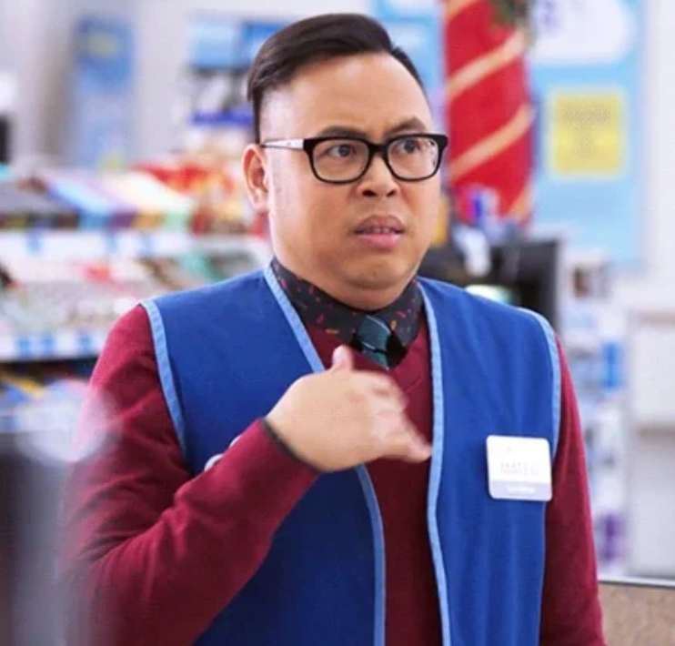 Retail worker in a store wearing a blue vest and name tag looks concerned or confused, touching their chest