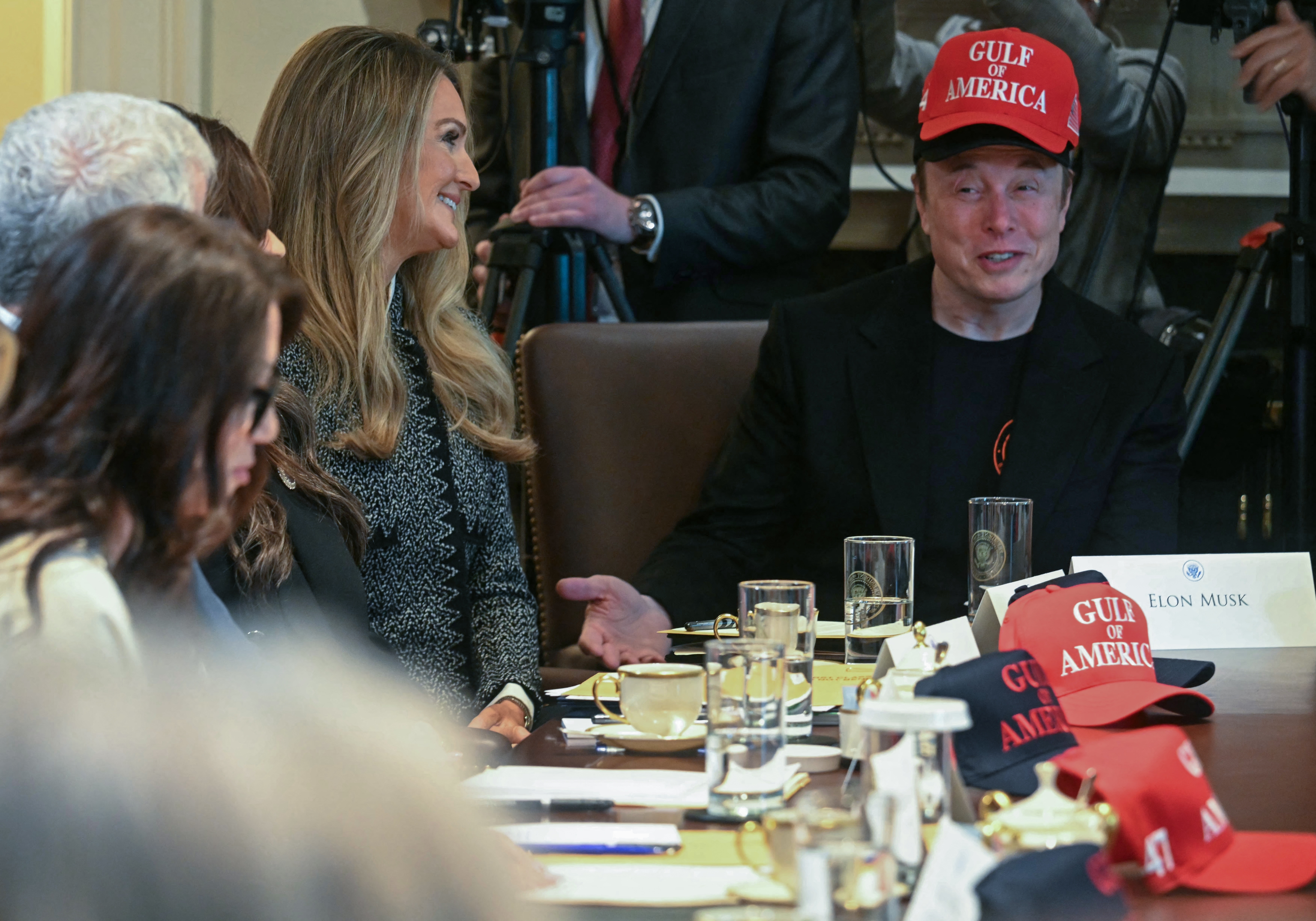 I don't know who is in the image, but it shows a meeting with several people seated around a table, one wearing a hat with text