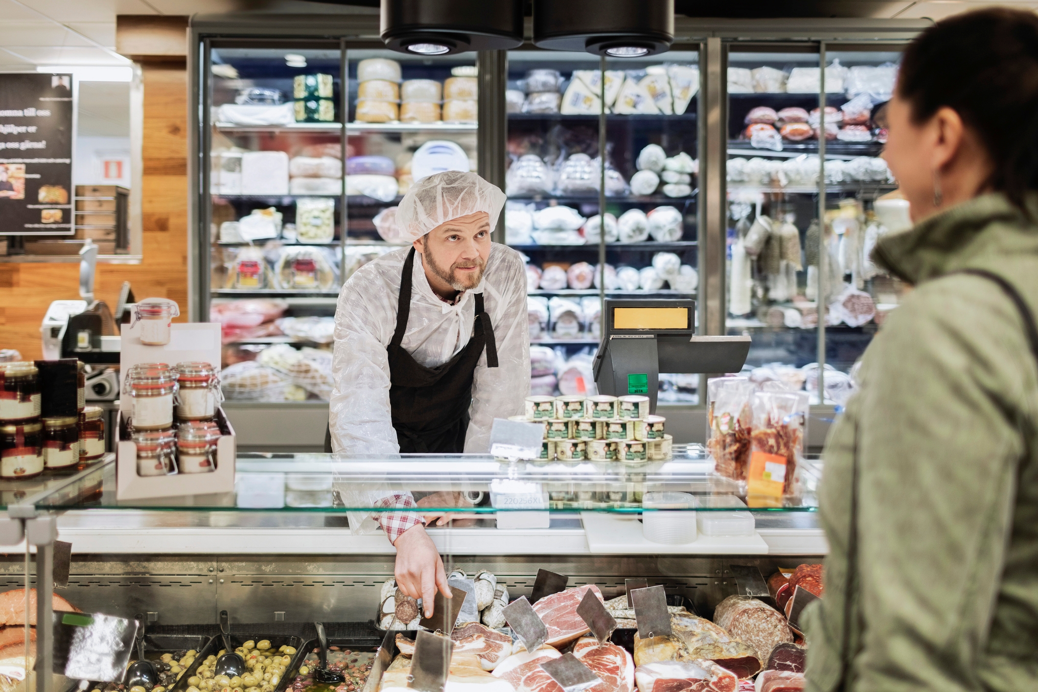 Store employee wearing a cap and apron assists a customer at a deli counter, surrounded by various meats and cheeses
