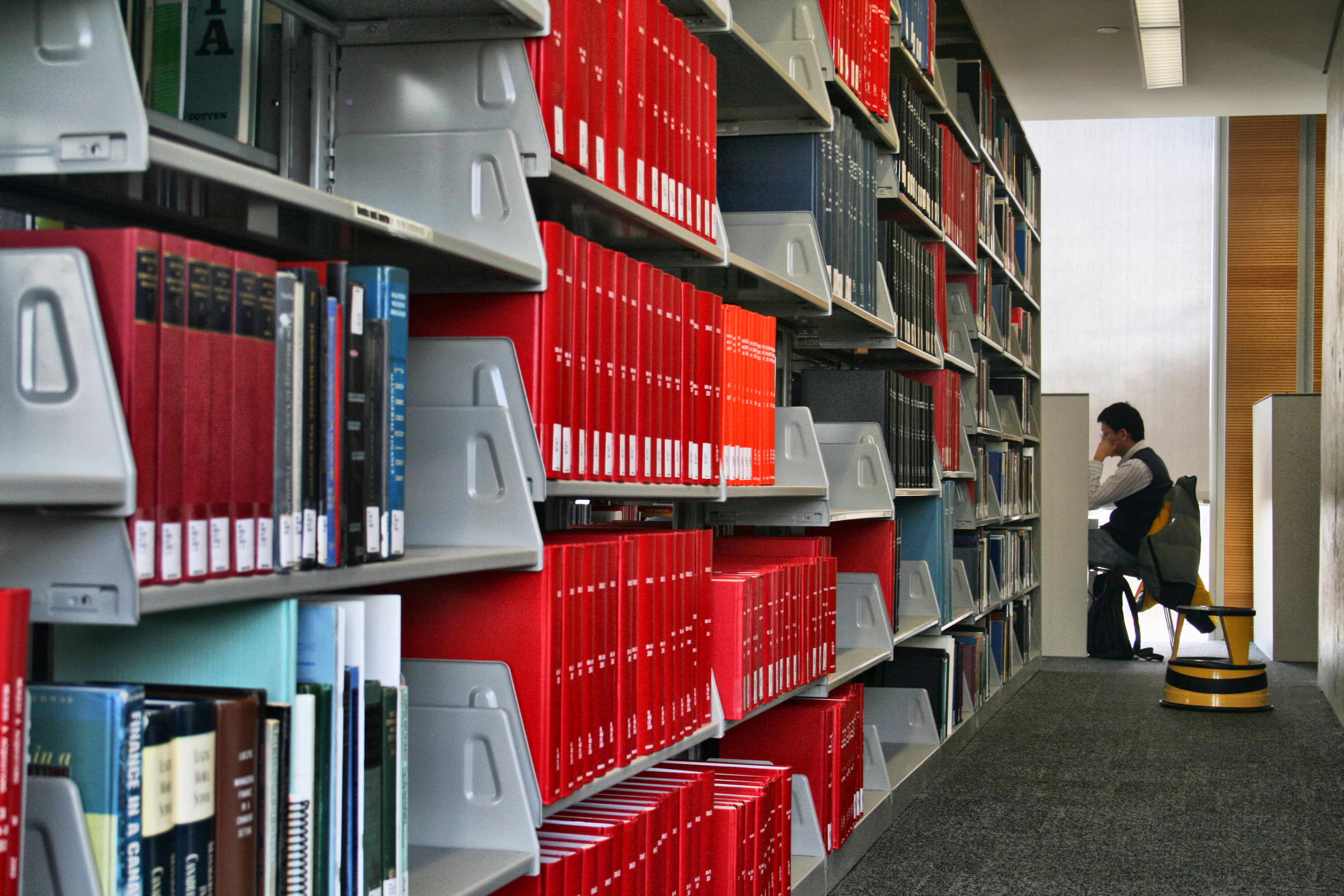 Person reading in library, seated at a desk. Shelves filled with books create a quiet, focused atmosphere