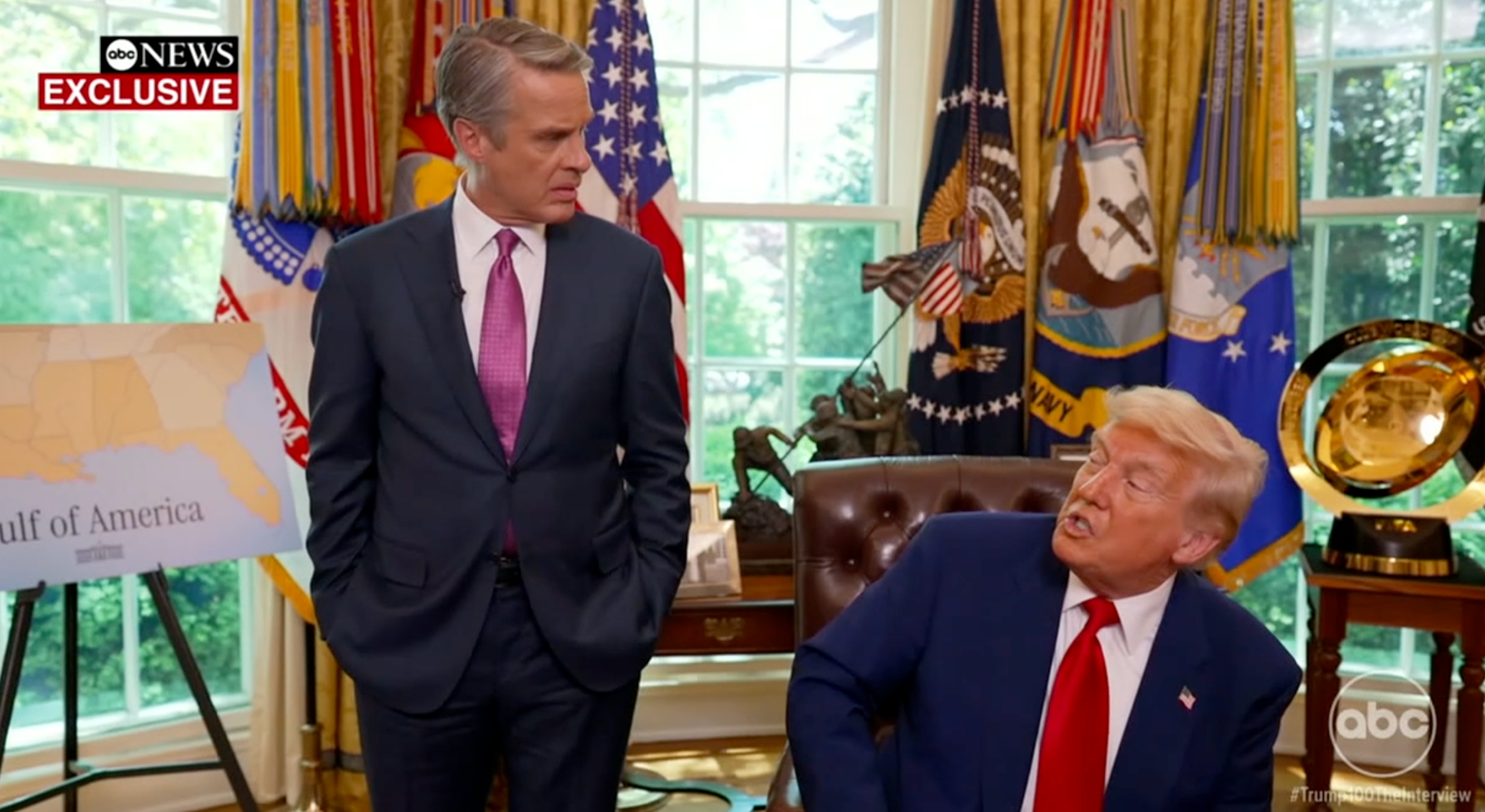 Two men in a formal office setting with flags and a map in the background. One is seated, wearing a suit, in conversation with the standing man