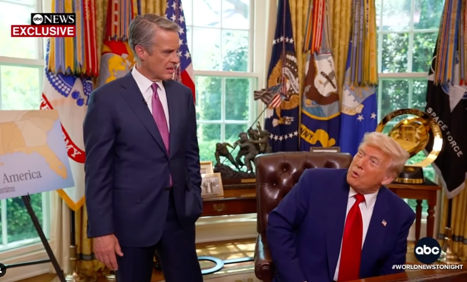 Two men in a formal setting with flags; one is seated at a desk, the other is standing. They appear engaged in conversation