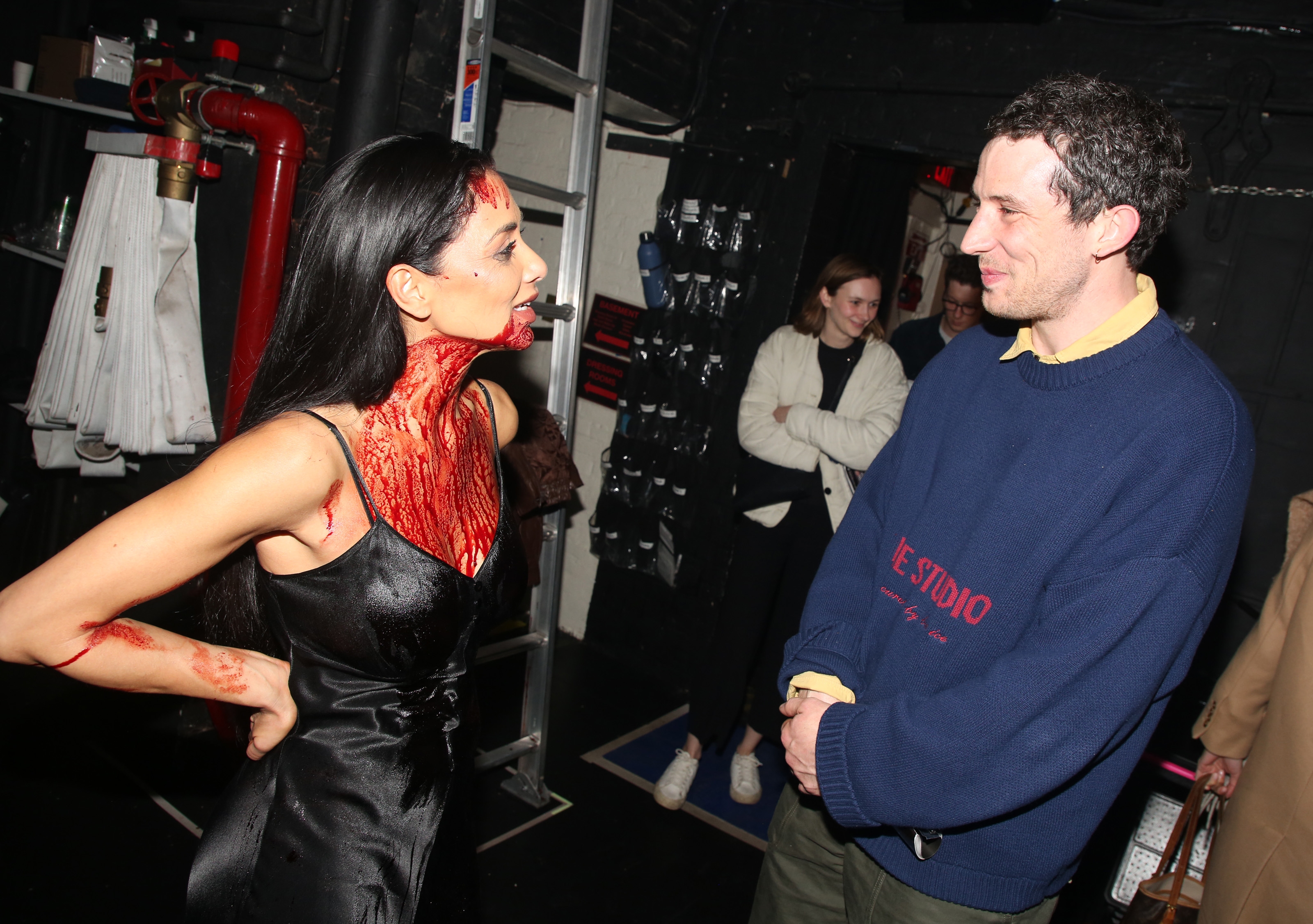 Person in theatrical makeup resembling blood converses with another backstage. They wear a sleek, sleeveless dress, while the other is in casual attire