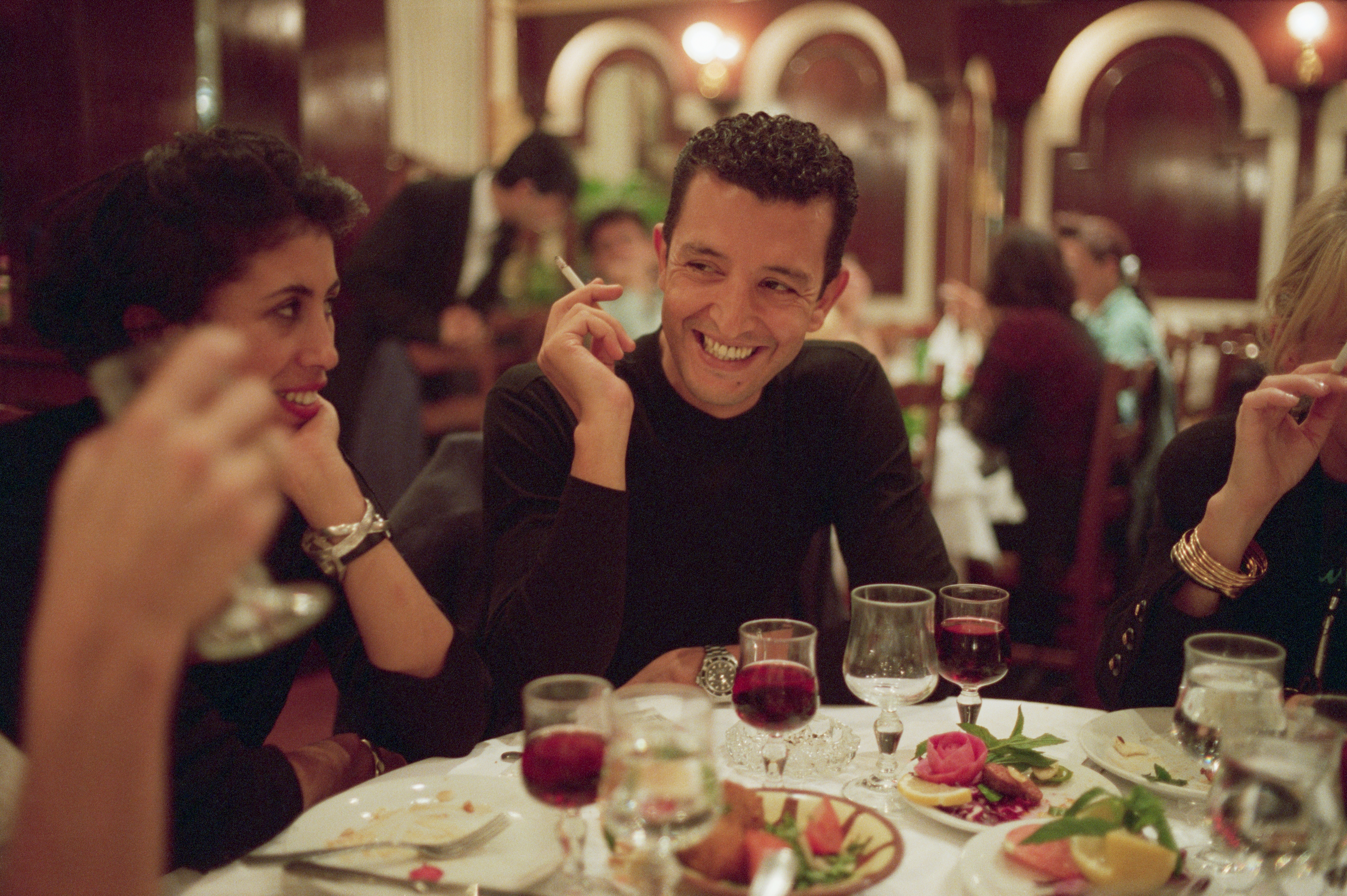 People dining, laughing, and conversing at a restaurant table with drinks and plates. The setting is lively and social
