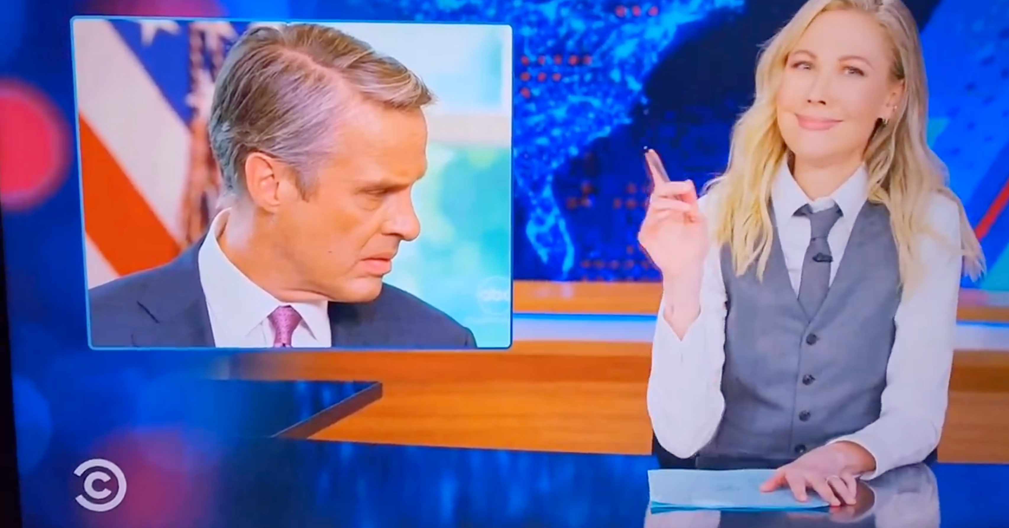 A TV host in a suit vest gestures toward an inset image of a serious-looking man in a suit. News-style set in the background