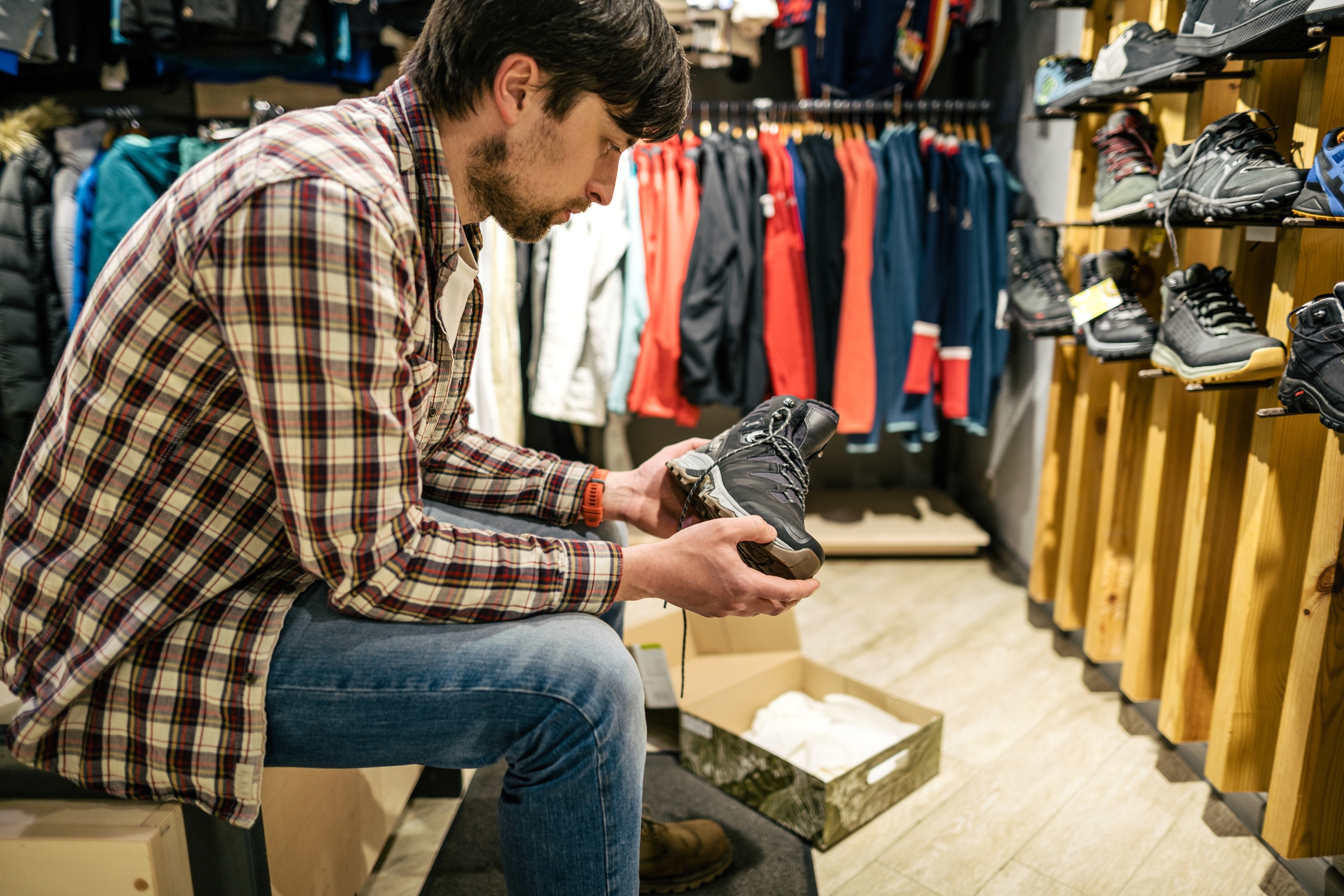 Man in plaid shirt examines sports shoes in a clothing store, seated by a shoe display, surrounded by hanging athletic wear