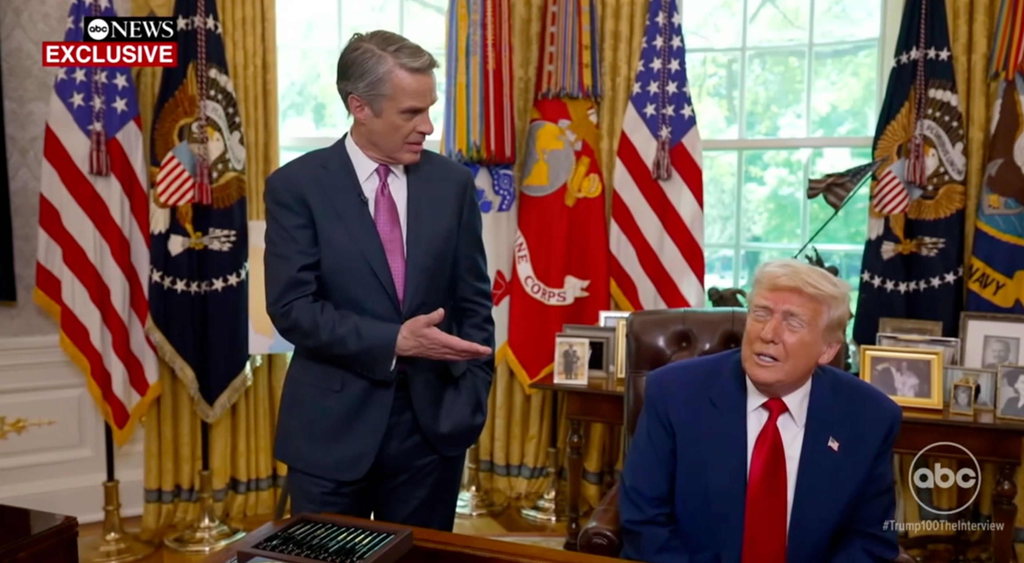 Two men in formal suits in an official-looking room, full of flags and framed photos; one is seated, and they appear to be having a discussion