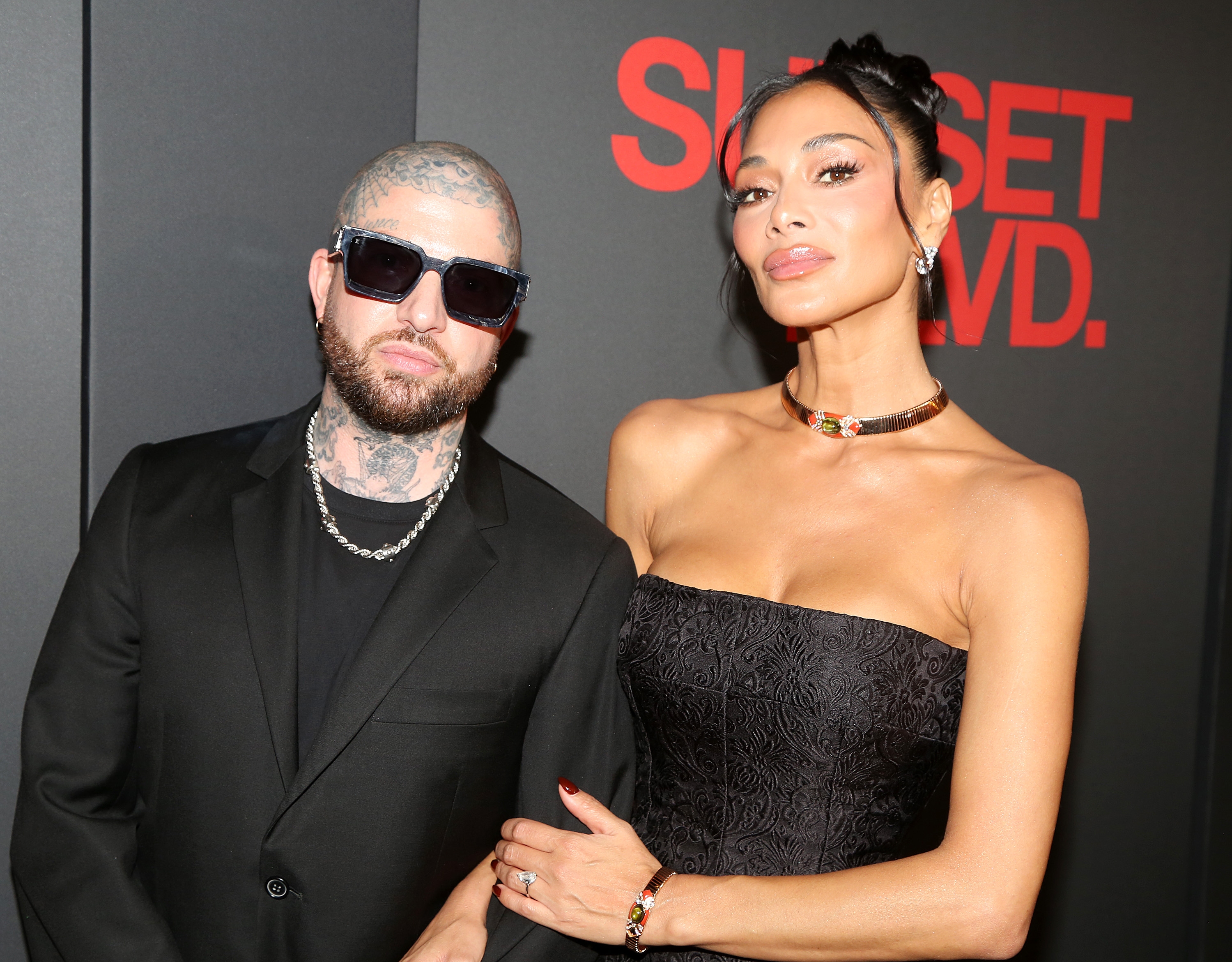 Two individuals pose on a red carpet. The person on the left wears a suit, sunglasses, and sports tattoos. The right person dons a strapless gown