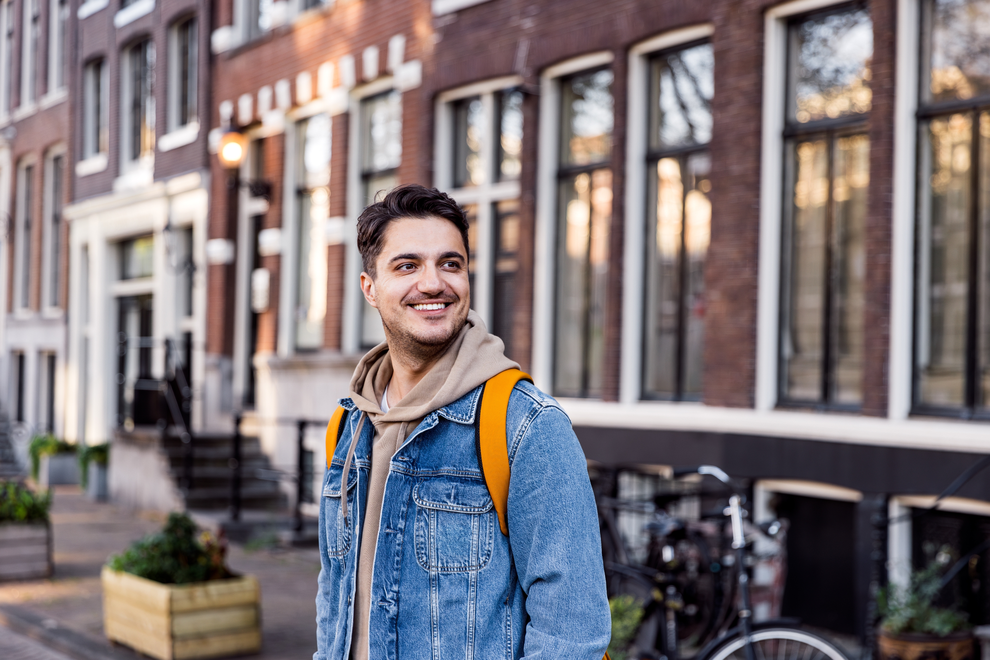 Person smiling outdoors, wearing a denim jacket and hoodie, standing on a city street with brick buildings