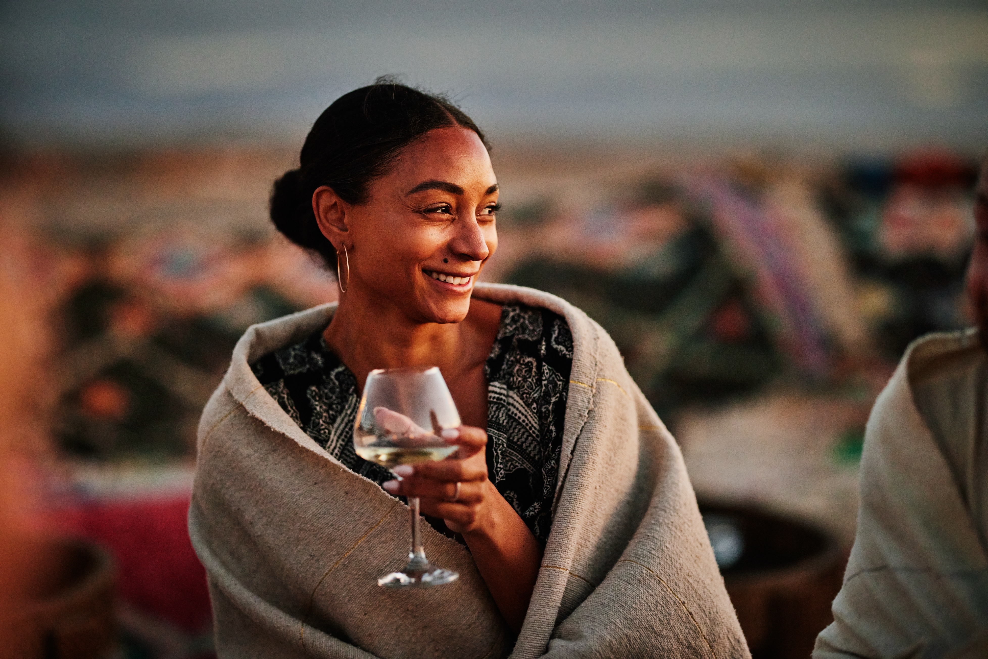 A woman wrapped in a blanket smiles while holding a wine glass outdoors. She is enjoying a relaxed moment in a cozy setting