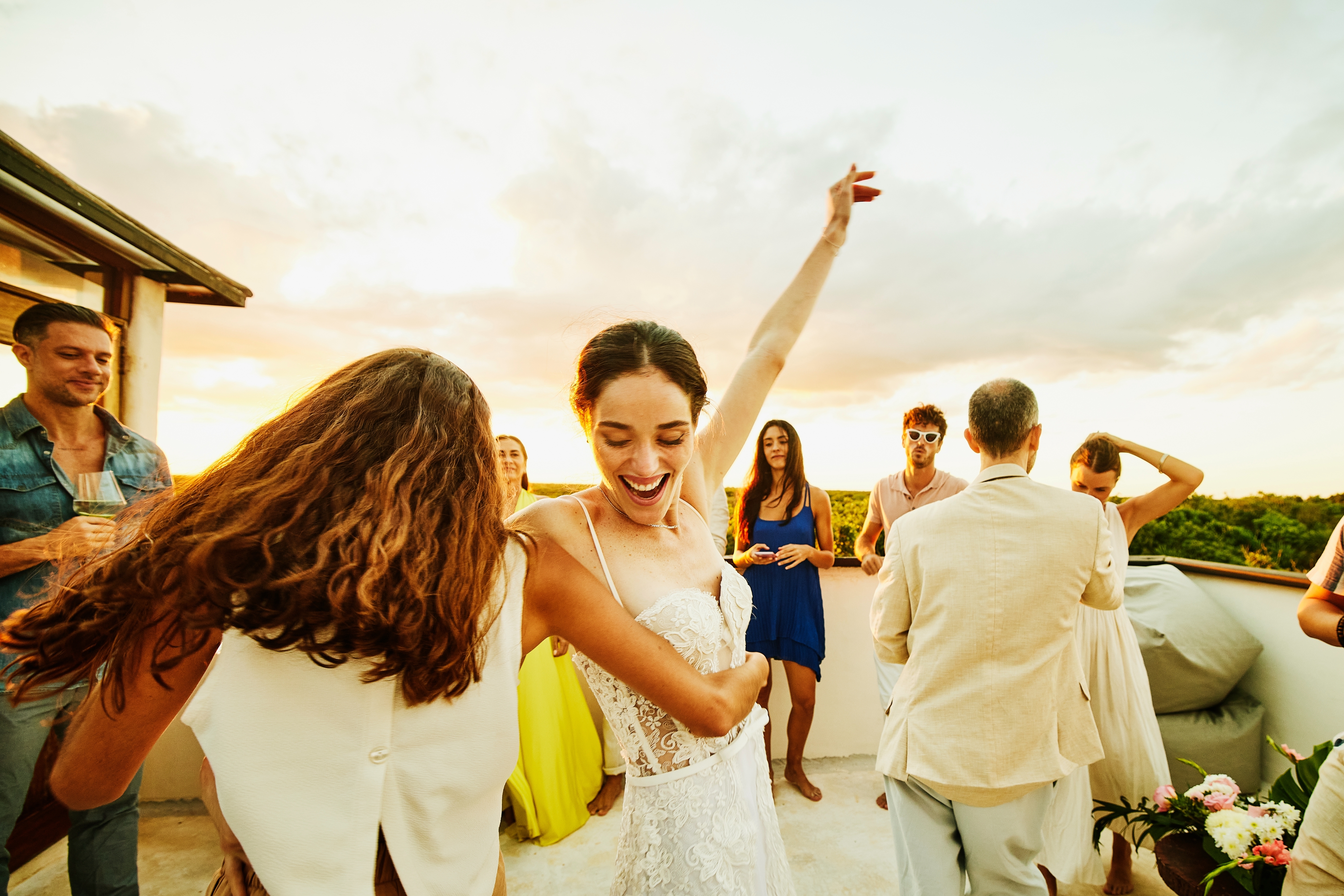 People dancing joyfully at an outdoor gathering during sunset, with one person in a lace dress raising an arm happily