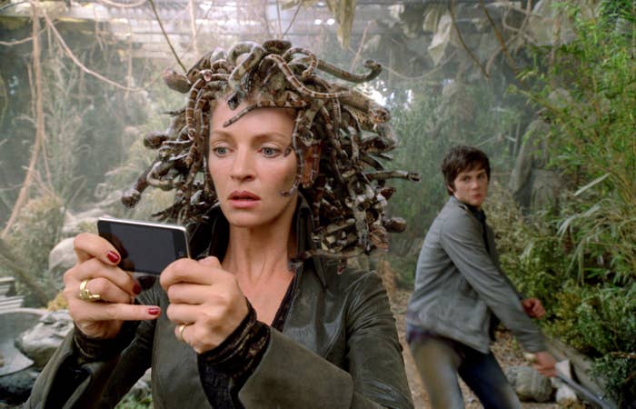 Woman with snake-like hair looks at smartphone; young man in a jacket appears in the background, looking away