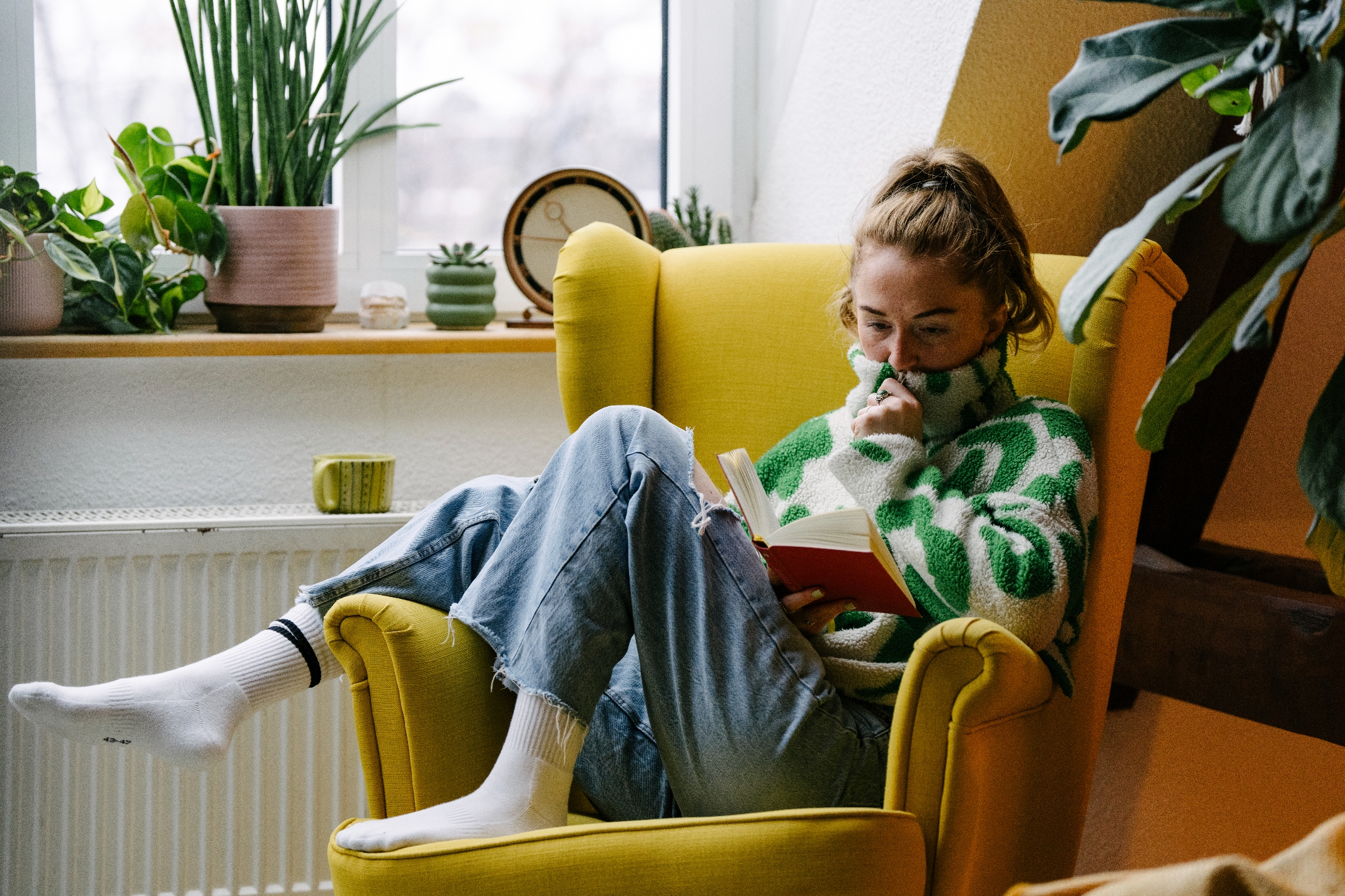 Person in cozy sweater reads on a yellow armchair, surrounded by potted plants in a bright room