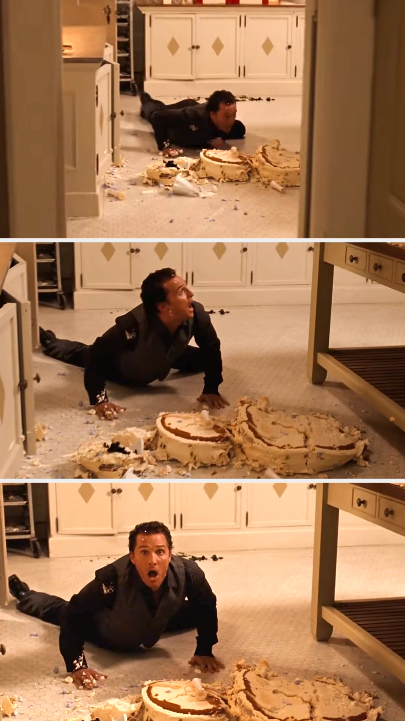 A man in a kitchen lies on the floor, surrounded by a large, collapsed cake, appearing surprised and concerned