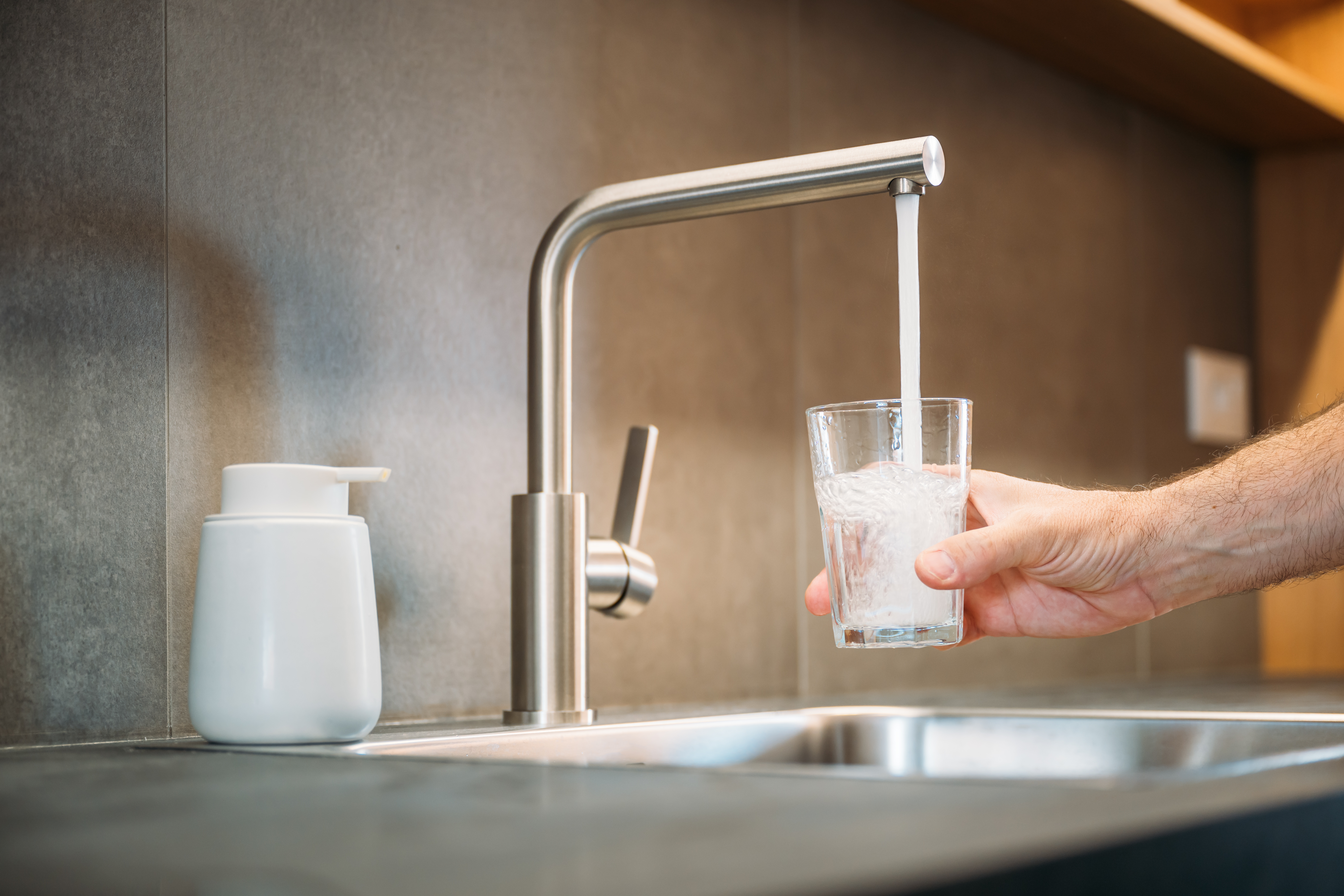 Hand holding a glass under a kitchen faucet, filling it with running water next to a soap dispenser