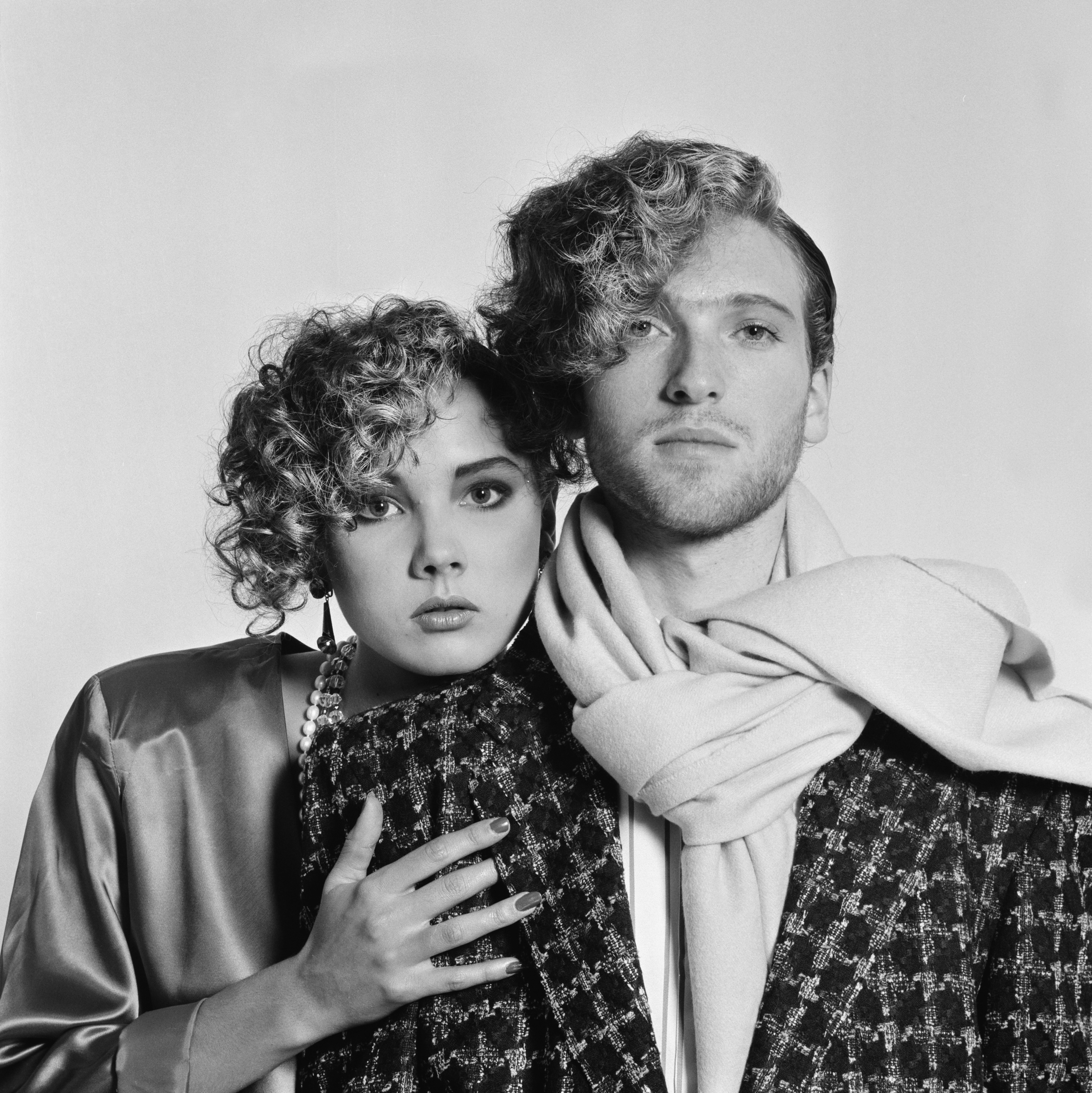 Two people with curly hair pose dramatically, wearing patterned outerwear and jewelry. The person on the right has a scarf draped around their neck
