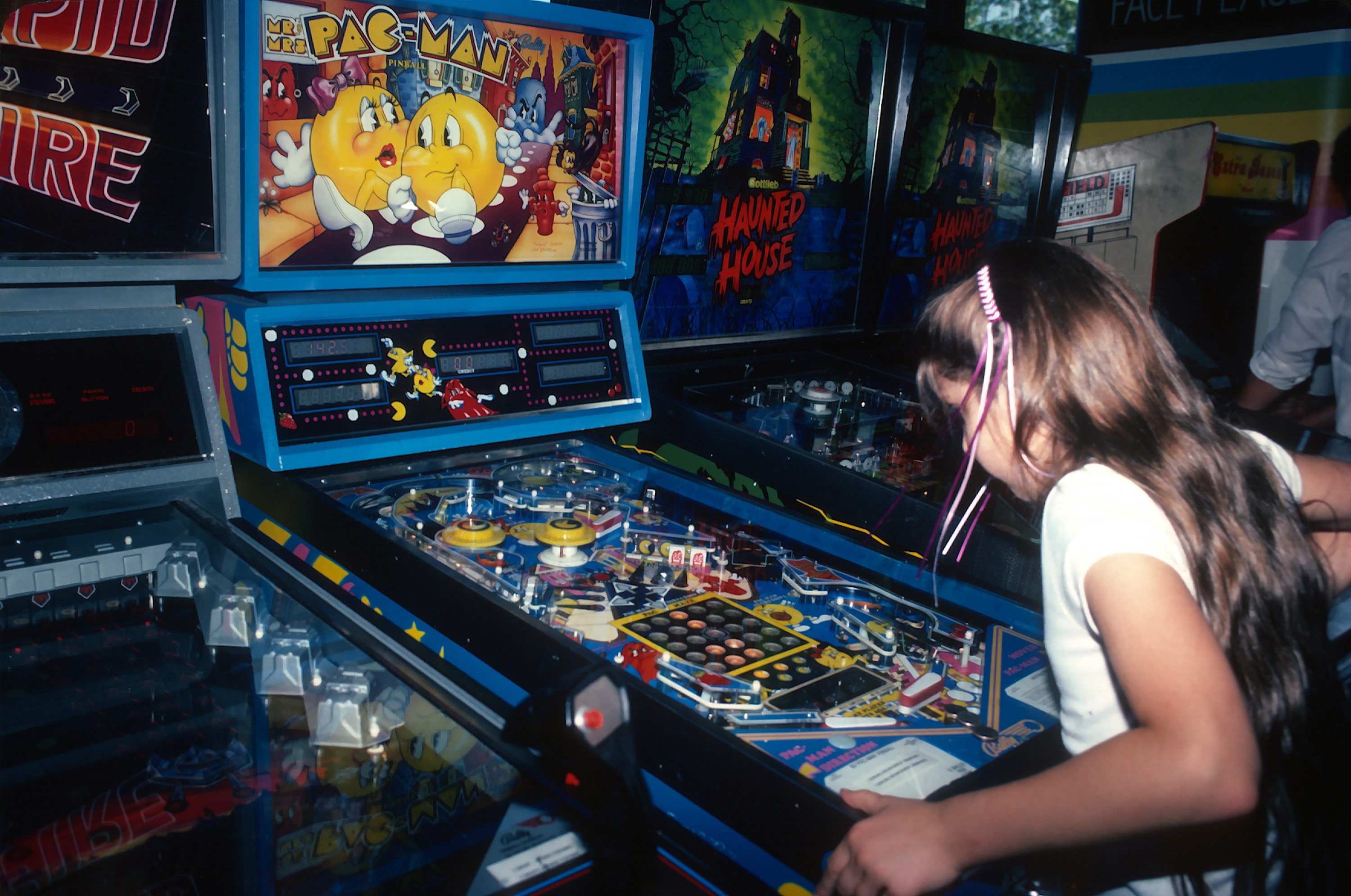 A young girl plays a vintage Pac-Man pinball machine in an arcade, focused on the game