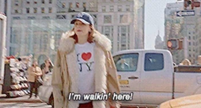 Person in a city street wearing a fur-trimmed coat, cap, and an "I ♥ NY" shirt, with the text "I'm walkin' here!" featured prominently