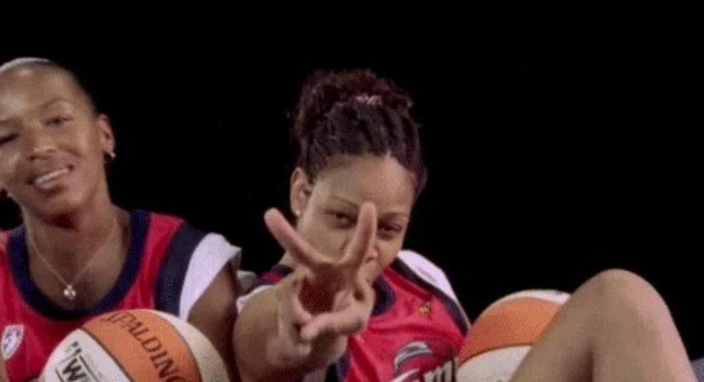 Two women in basketball jerseys sit with basketballs; one playfully gestures a peace sign towards the camera