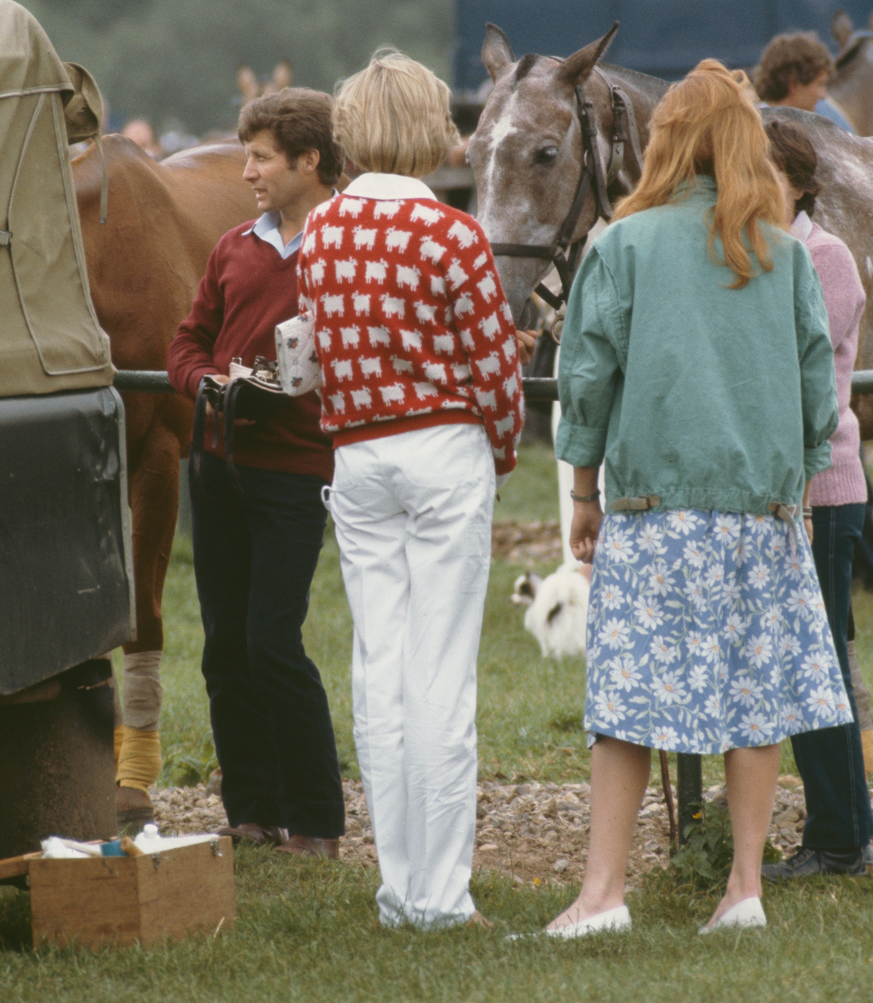 People at an outdoor event, one wearing a red sweater with white figures and the other in a floral skirt. Horses are in the background