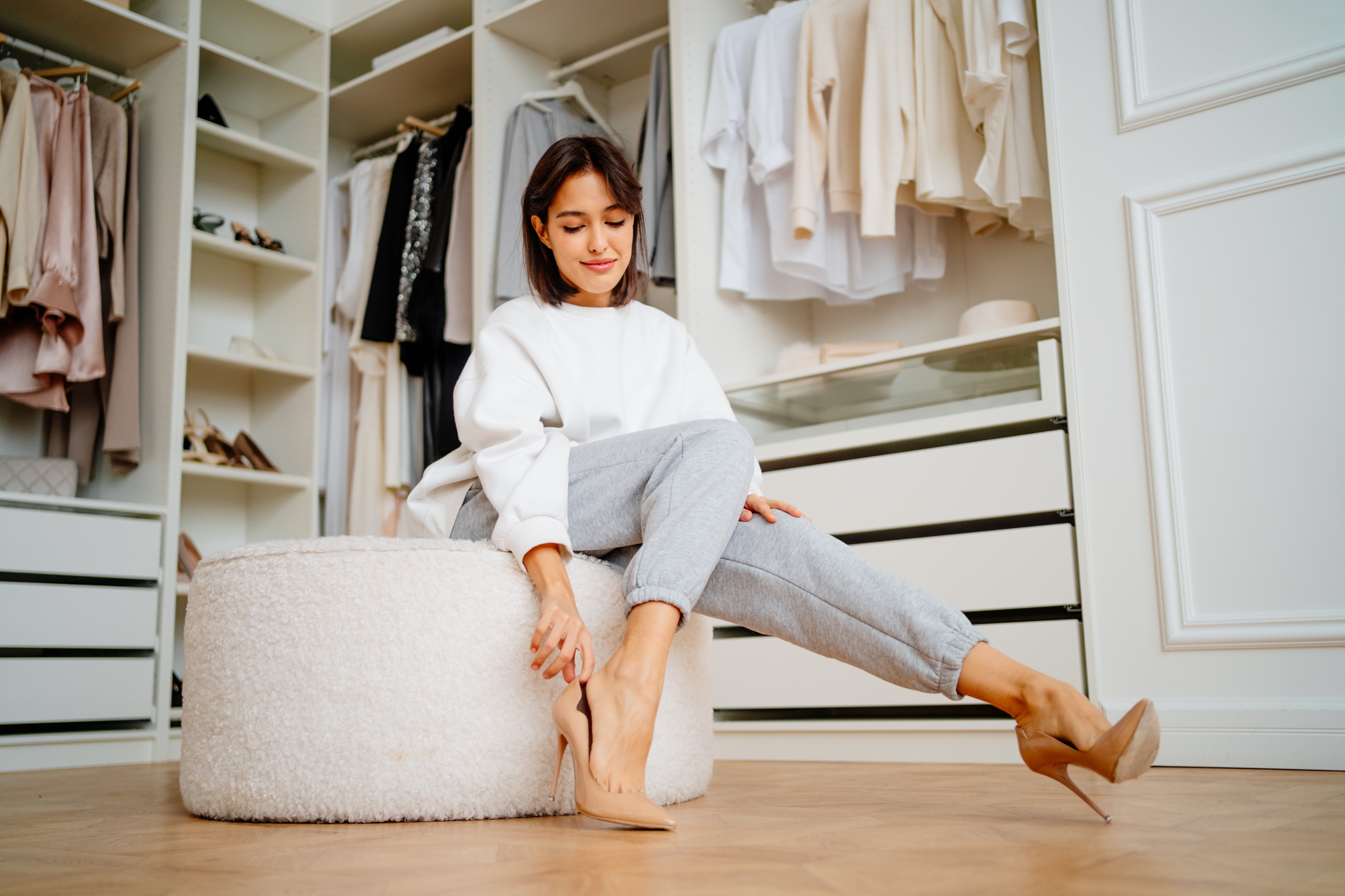 A woman in casual clothing, seated in a closet, is trying on high heels. Shelves of clothes and shoes are visible in the background