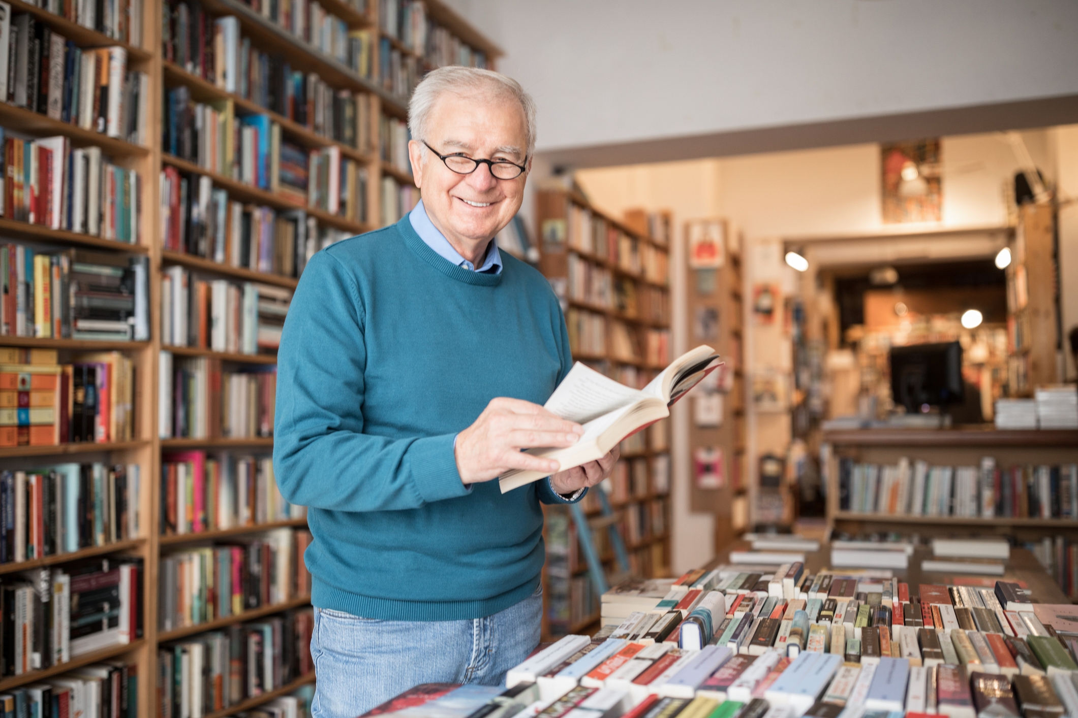An elderly man smiles while standing in a bookstore, holding an open book. Shelves filled with books surround him