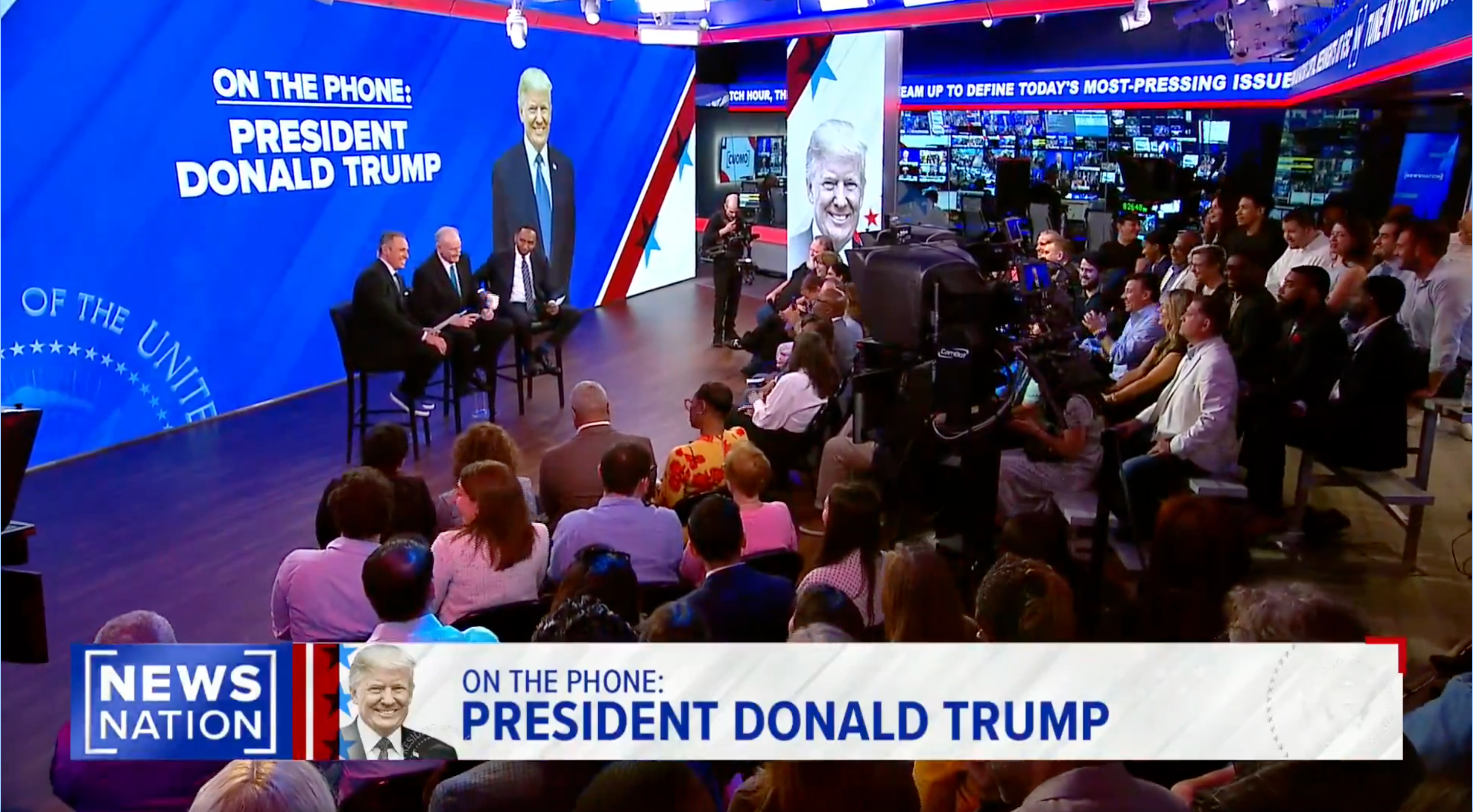 News studio audience laughs while watching a broadcast with Donald Trump joining via phone call
