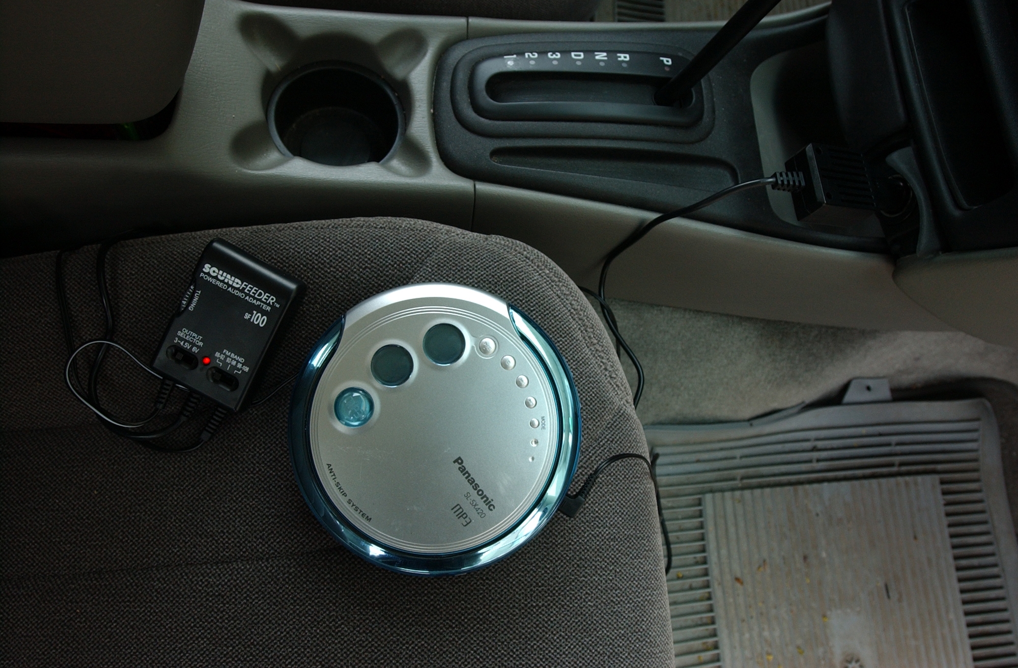 Portable CD player connected to a car stereo via cassette adapter on a car seat. Gearshift and cup holder are visible nearby