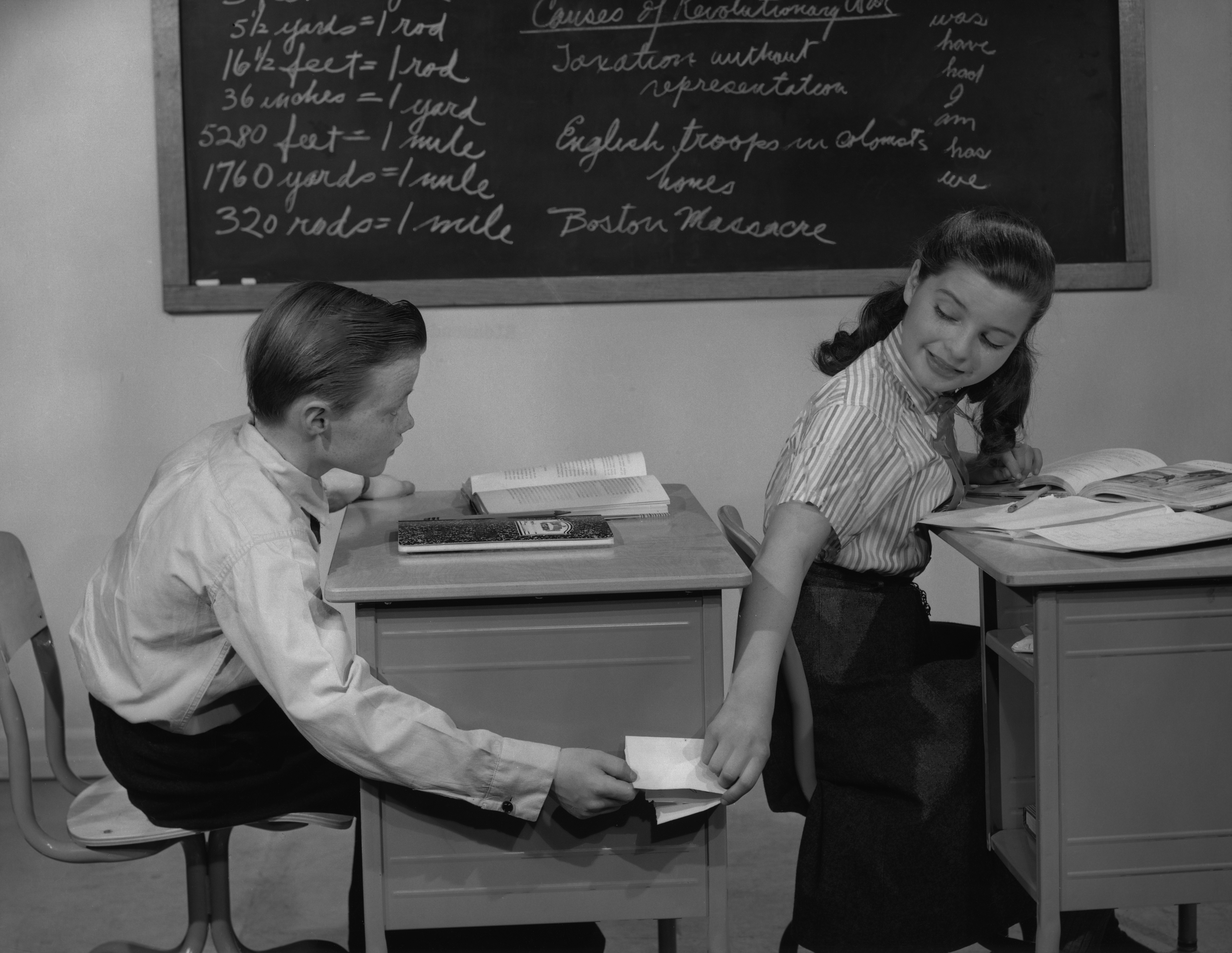 Student passing a note to a classmate behind a desk in a classroom with a chalkboard displaying math and geography topics