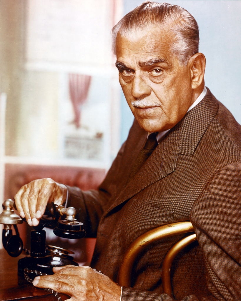 An older man with a mustache in a suit looks at the camera while seated, holding a vintage rotary telephone