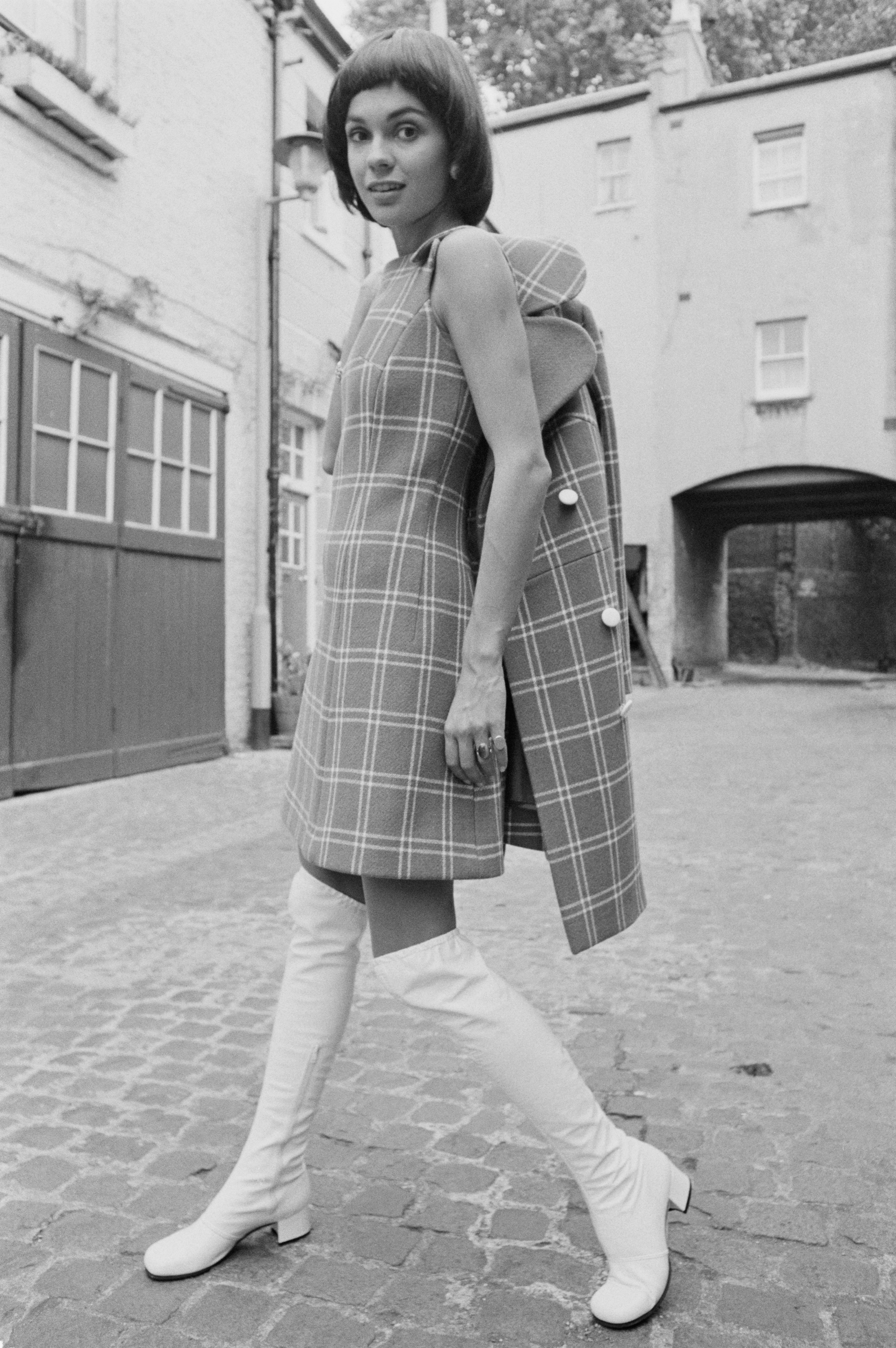 Woman in a checkered dress with matching coat and knee-high boots walking on a cobblestone street