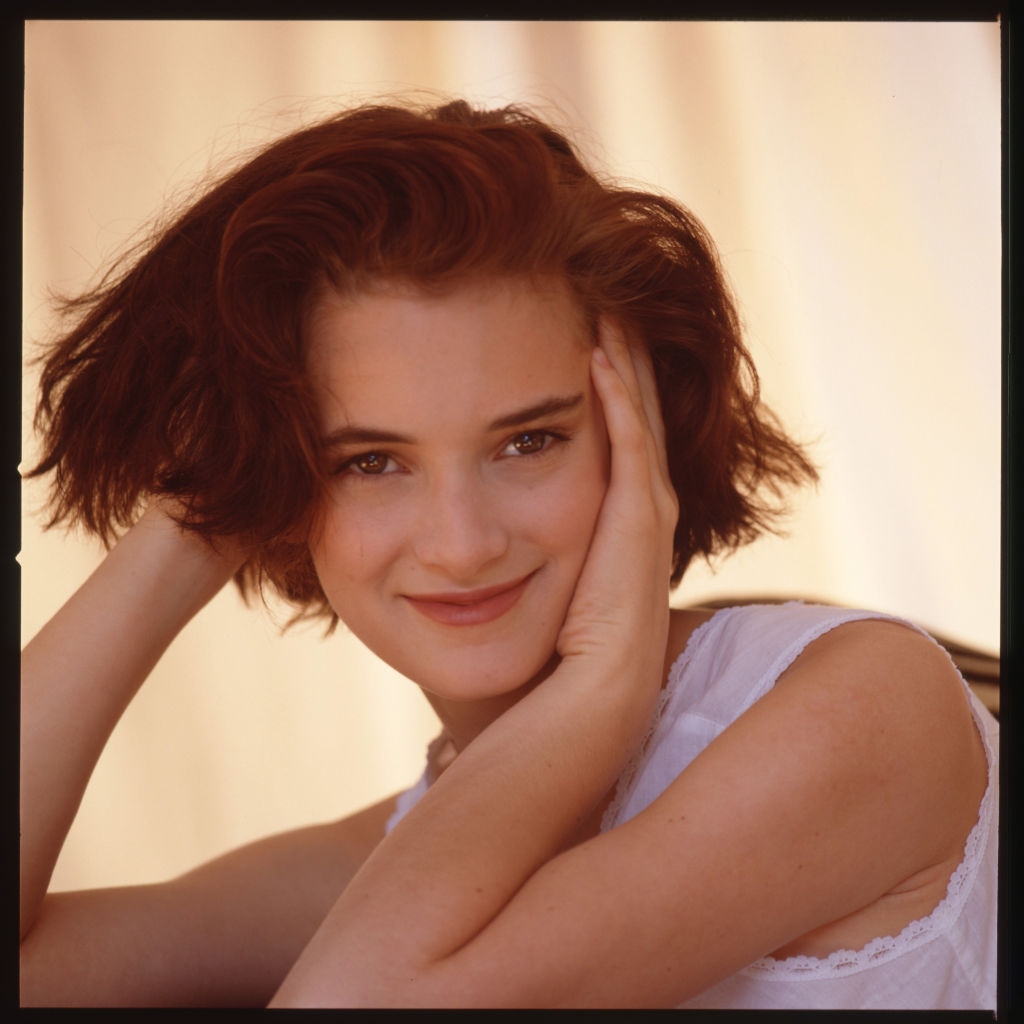 A person with short hair gently resting their head on their hands, smiling at the camera, wearing a lace-trimmed attire