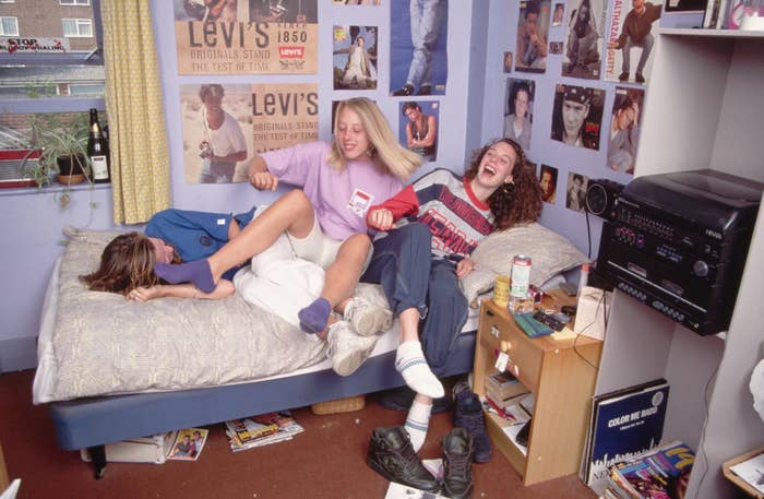 Three individuals laughing on a bed in a poster-filled room. One person is lying down, and two are sitting, playfully engaging with each other