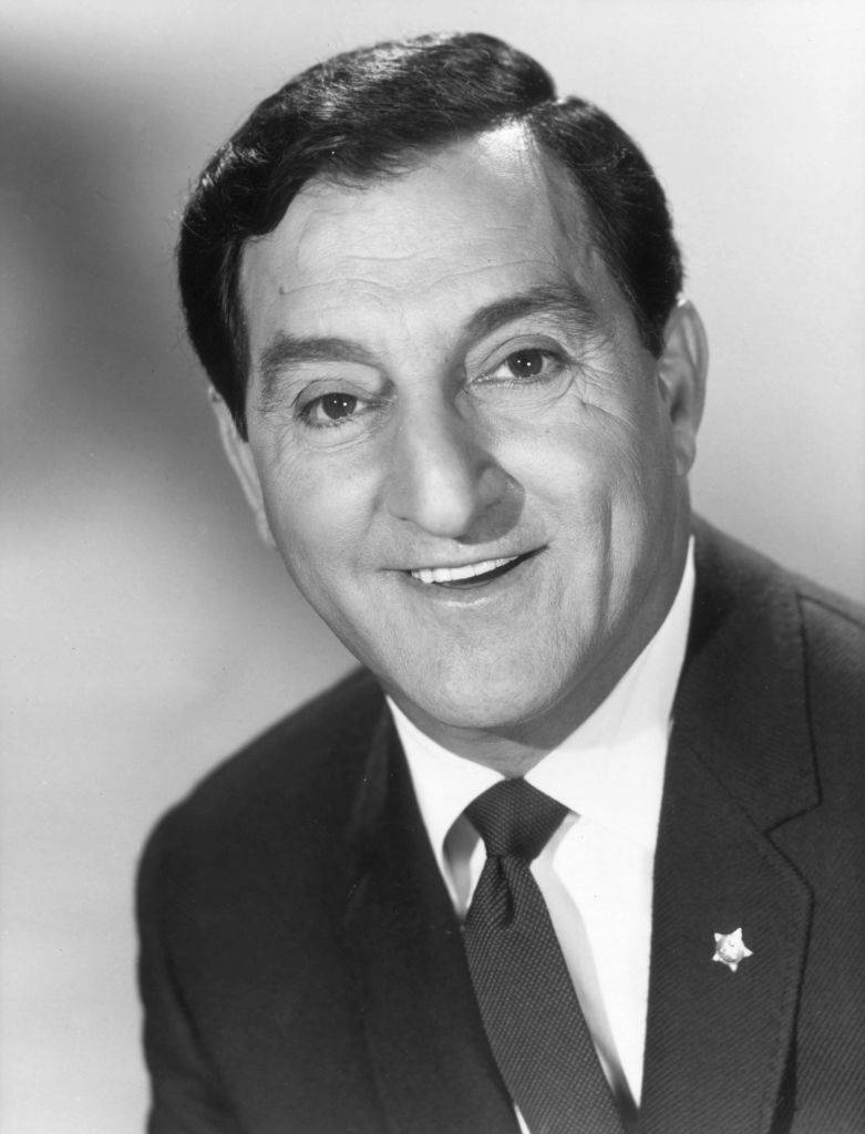 Man in suit and tie smiling, wearing a lapel pin, facing the camera