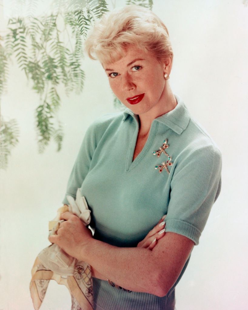 Person in a collared sweater with retro hairstyle and brooch, holding a scarf, poses in front of leafy background