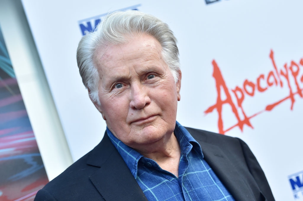 Person wearing a blue plaid shirt and dark blazer at an event with &quot;Apocalypse&quot; text on backdrop