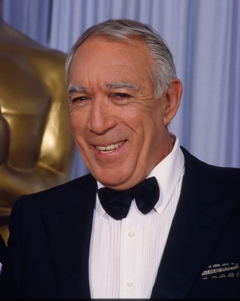 Smiling man in a tuxedo stands in front of an Oscar statue at an awards event