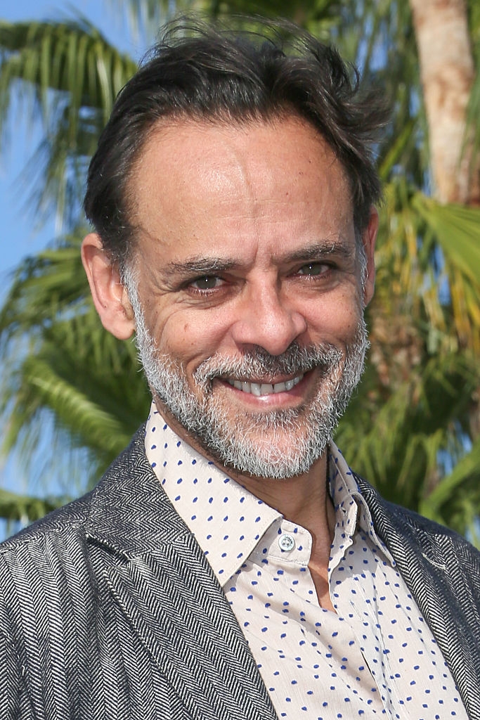 Smiling man with short hair and beard, wearing a patterned shirt and textured jacket. Palm trees in the background