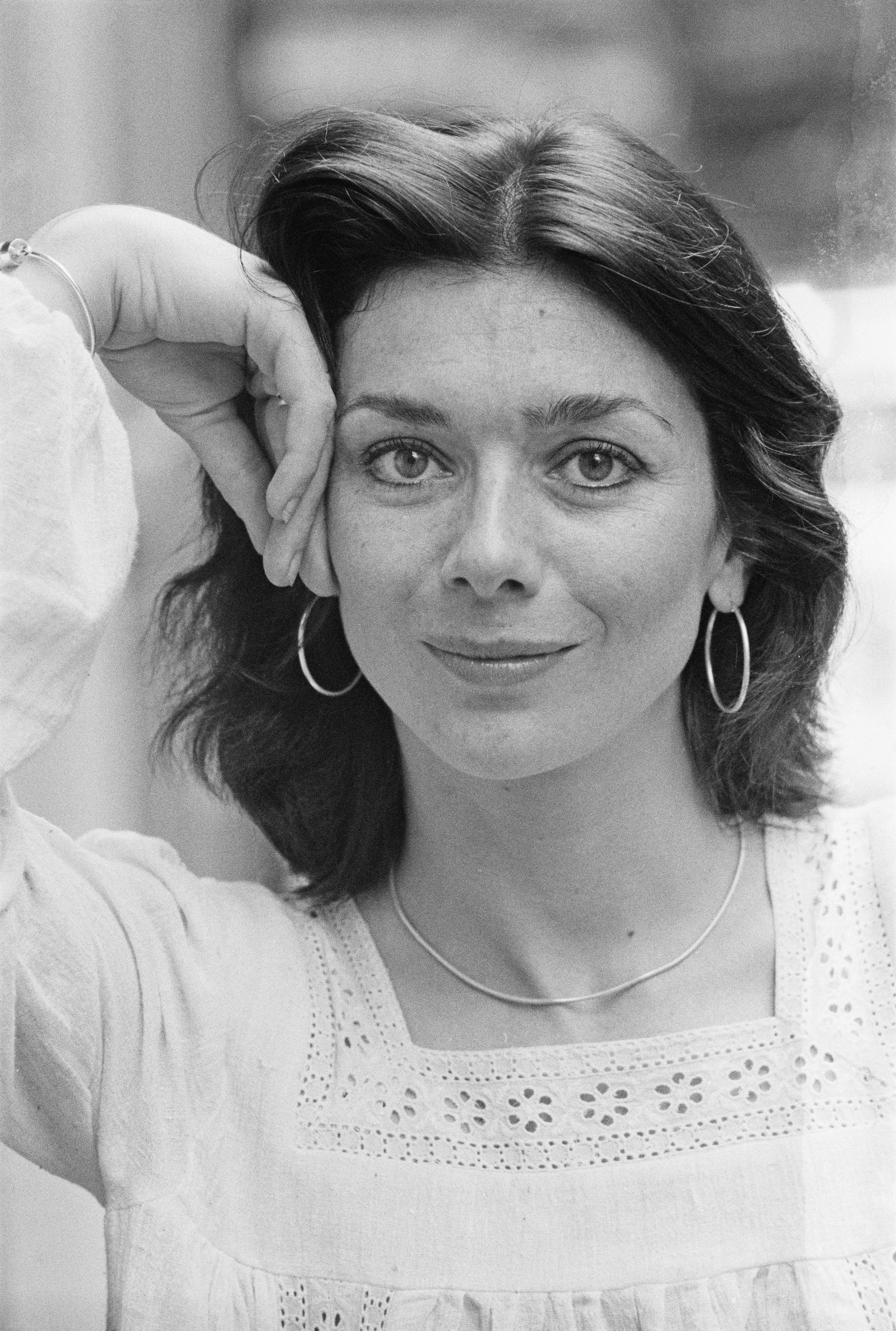 Person with hoop earrings and a delicate necklace, wearing a detailed embroidered top, rests their head on their hand, smiling softly at the camera