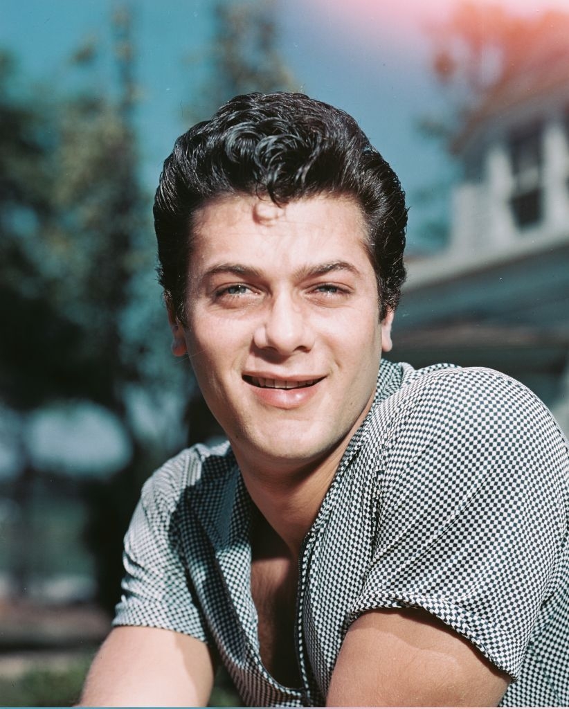 Person outdoors, smiling, wearing a checkered shirt. Trees and a building in the background