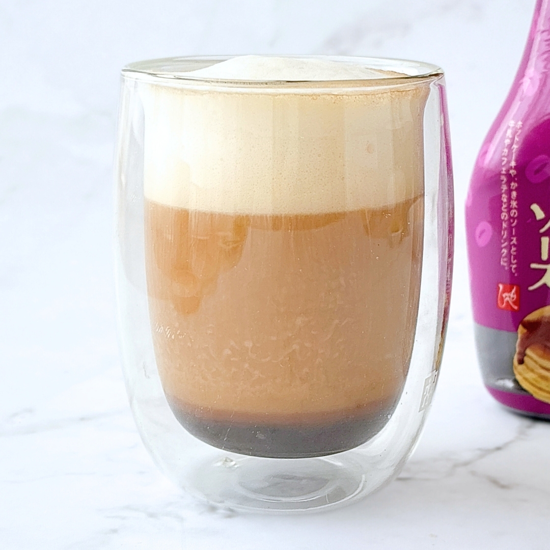 クリーミーなトップのアイスコーヒーが透明なガラスコップに入っている。隣に赤色の包装が見える。