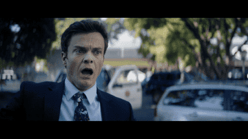 口を開けた驚いた表情の男性が、外で車や木を背景に映っている。