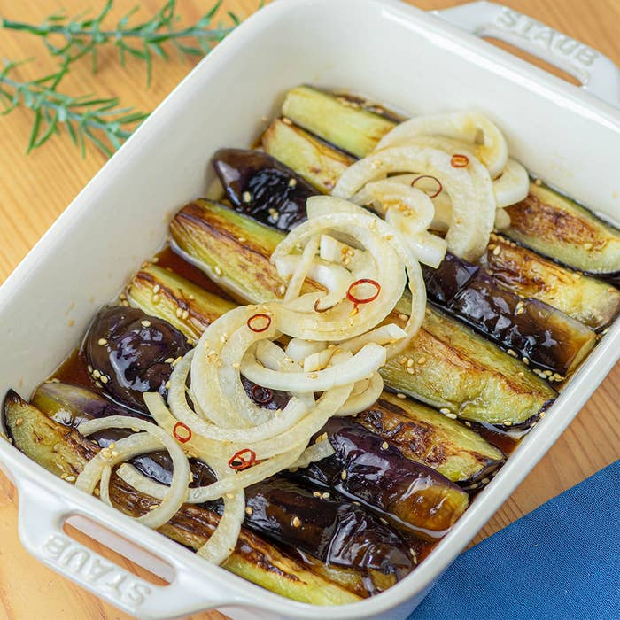 焼きナスの上にタマネギと唐辛子のスライスがのった白いキャセロール皿。テーブルに青い布が置かれている。