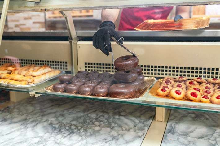 ディスプレイケースの中で店員がチョコレートドーナツをトングで取っている場面。周りには他のペストリーも並んでいる。