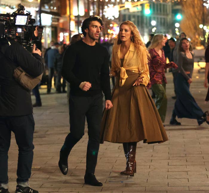 A man and woman walk on a busy street; the man is in casual attire, and the woman in a vintage-style blouse and skirt, with film crew nearby