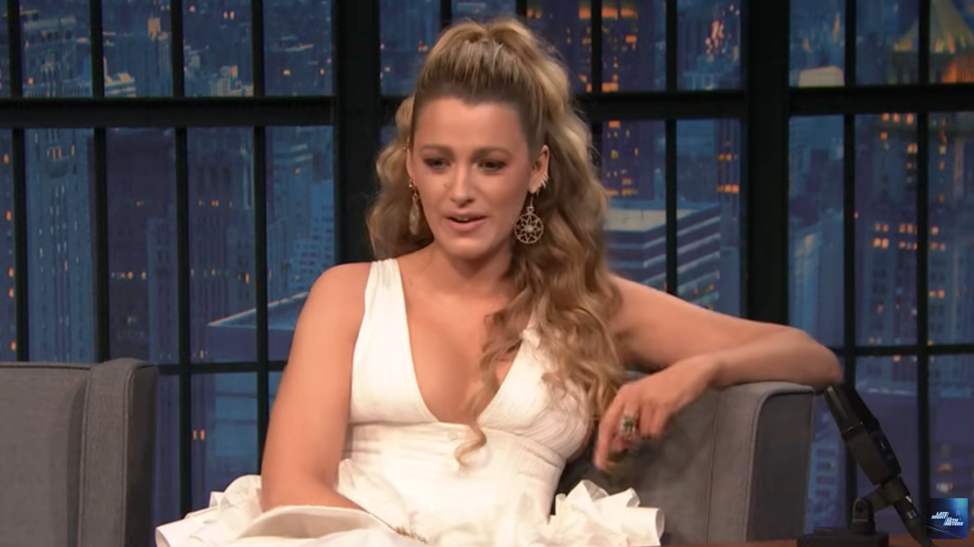 Person in an elegant dress sits on a talk show set, engaging in conversation, with a cityscape visible in the background